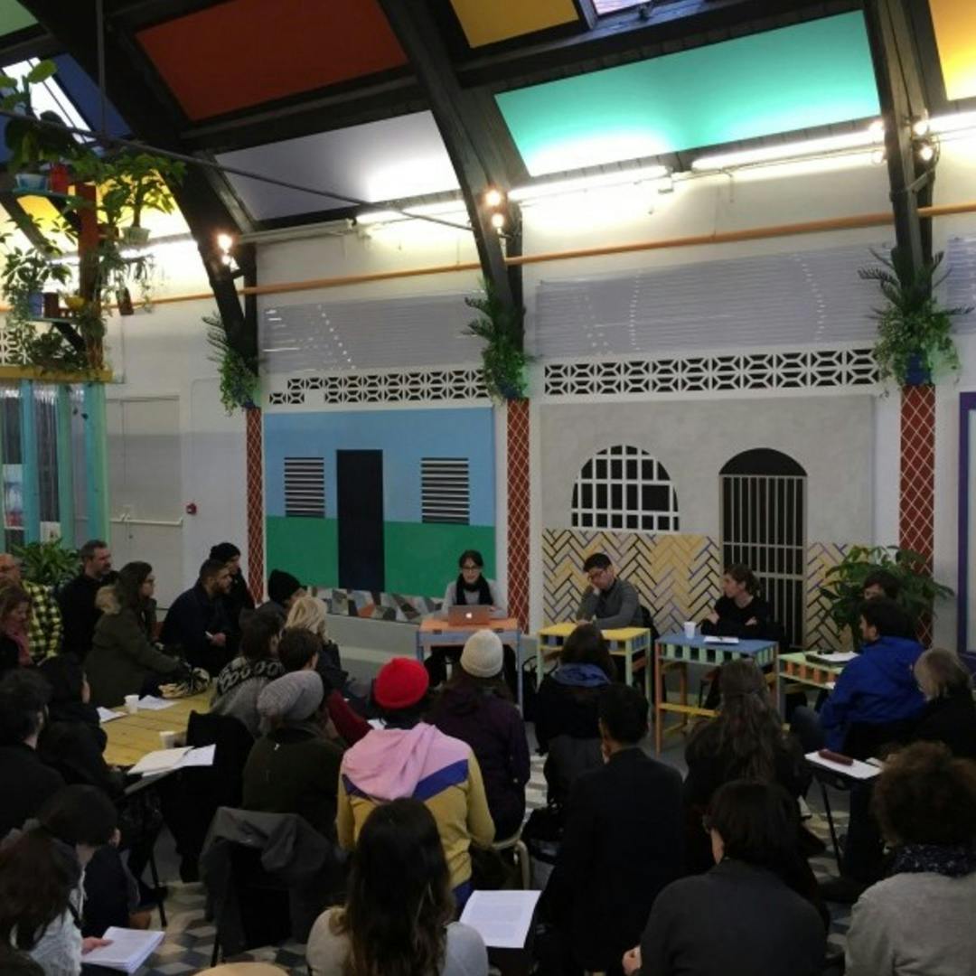 A group of people sit in a circle on stools inside a colourful, decorated room, attentively listening to a panel of speakers at the front. Some participants hold papers, and the atmosphere appears collaborative and engaged.