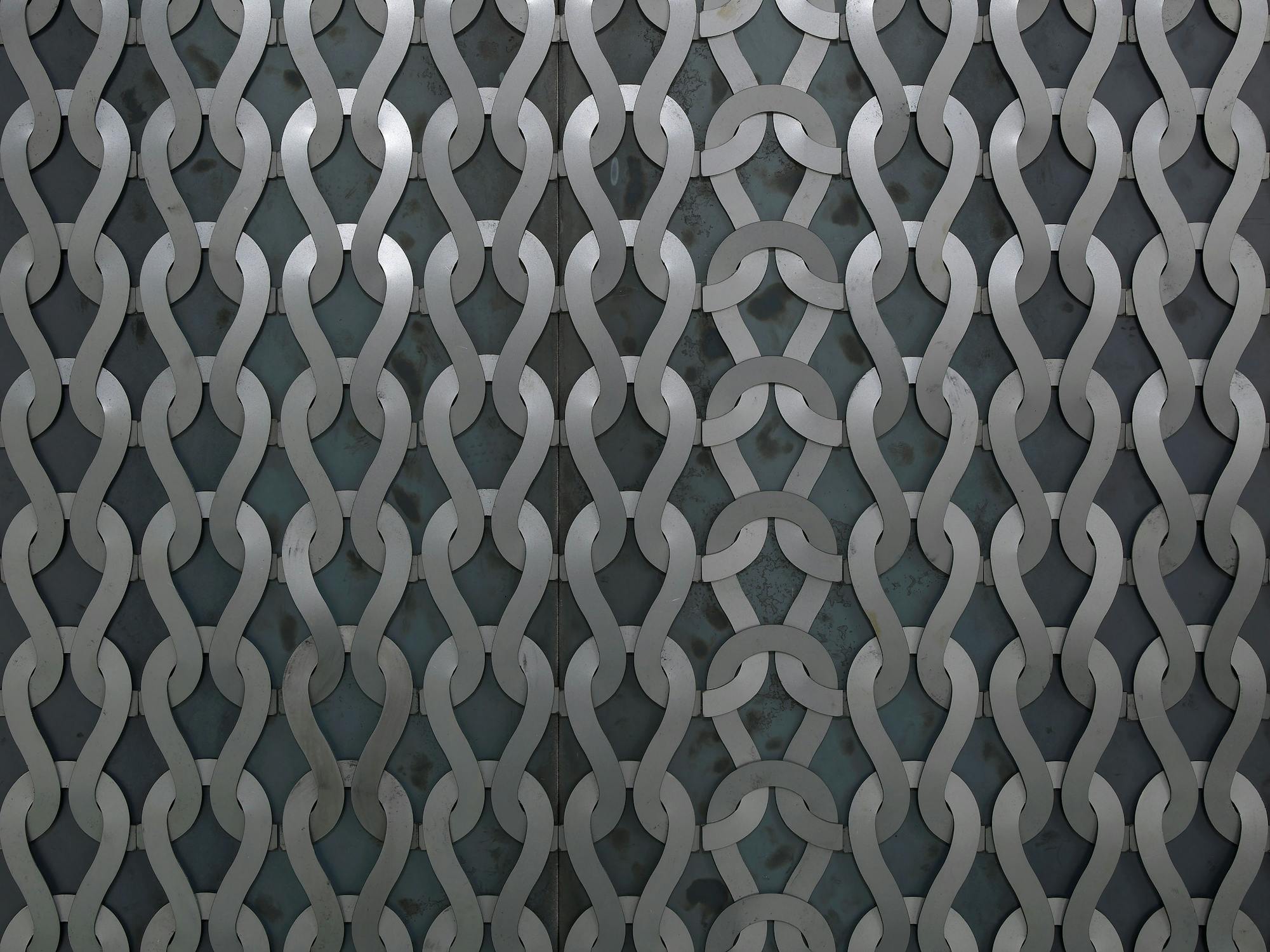 A close-up of an interwoven metal grid with a repeating pattern of silver, curved strips forming a chain-like design over a dark, textured background. The metal strips create a sense of depth and geometric complexity.