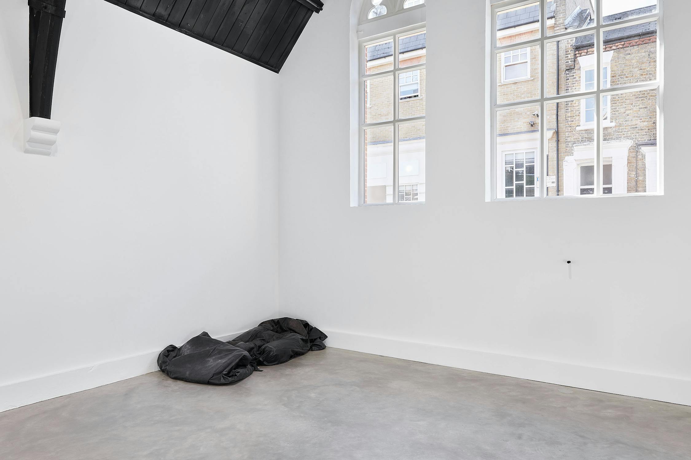 A bright, minimal room with white walls, a concrete floor, large windows. A black leather bag, resembling a body bag in the corner. The windows show brick buildings outside.