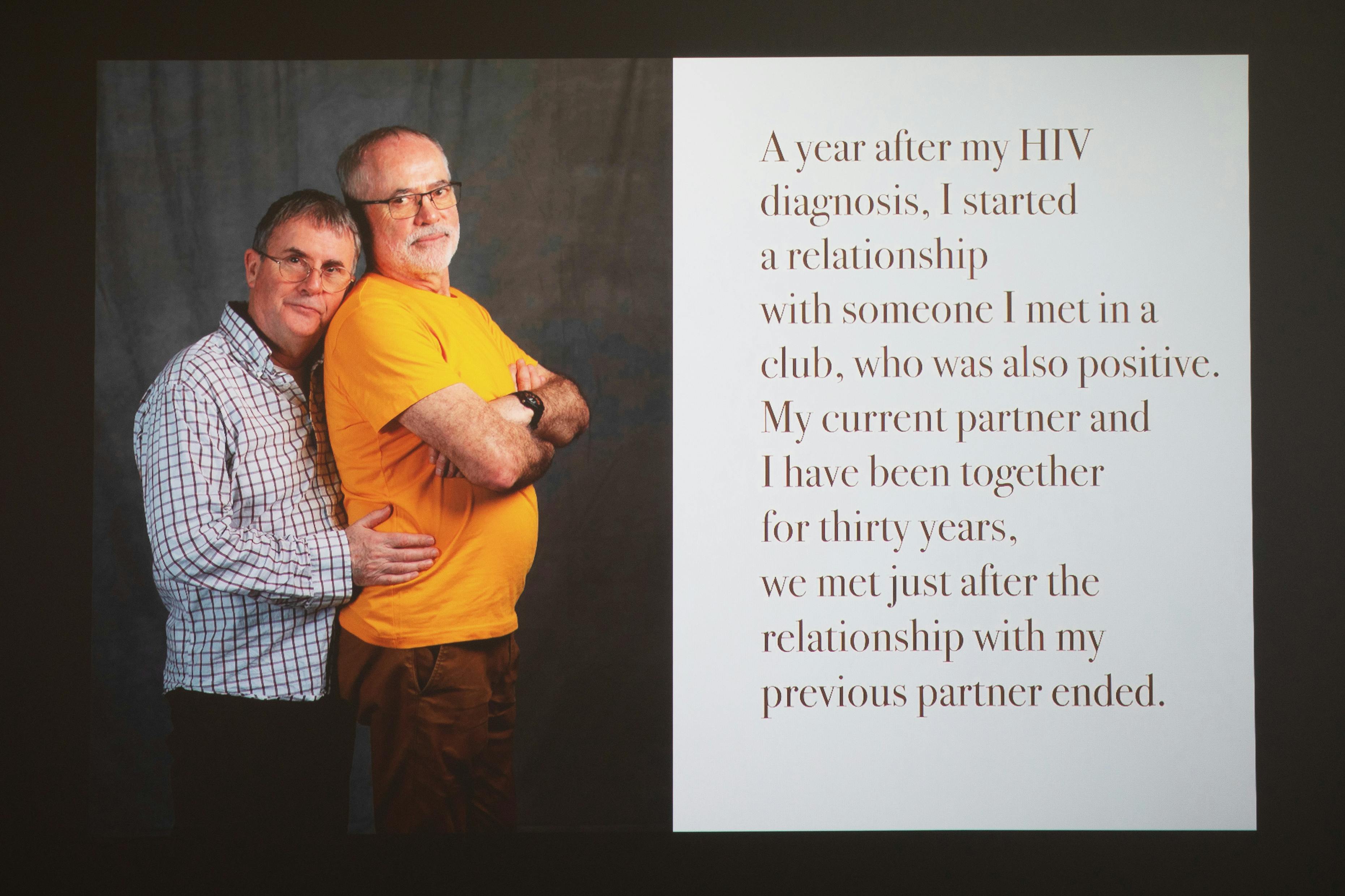 A photo of two men standing close together, one in an orange shirt with his arms crossed, and the other in a plaid shirt with his arm around the first man's waist. Beside them is a text detailing their relationship and living with HIV for over thirty years.