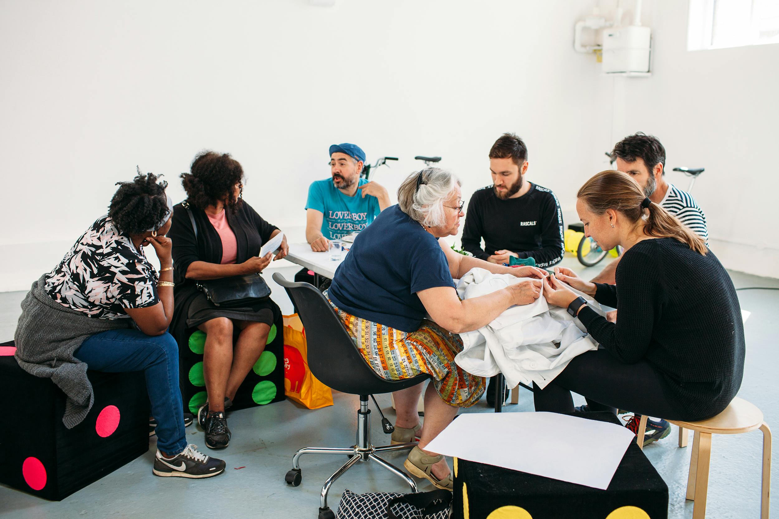 A diverse group of people sit in a circle on stools and colourful cube seats, engaged in conversation and activities, in a bright, modern room with white walls and natural light.