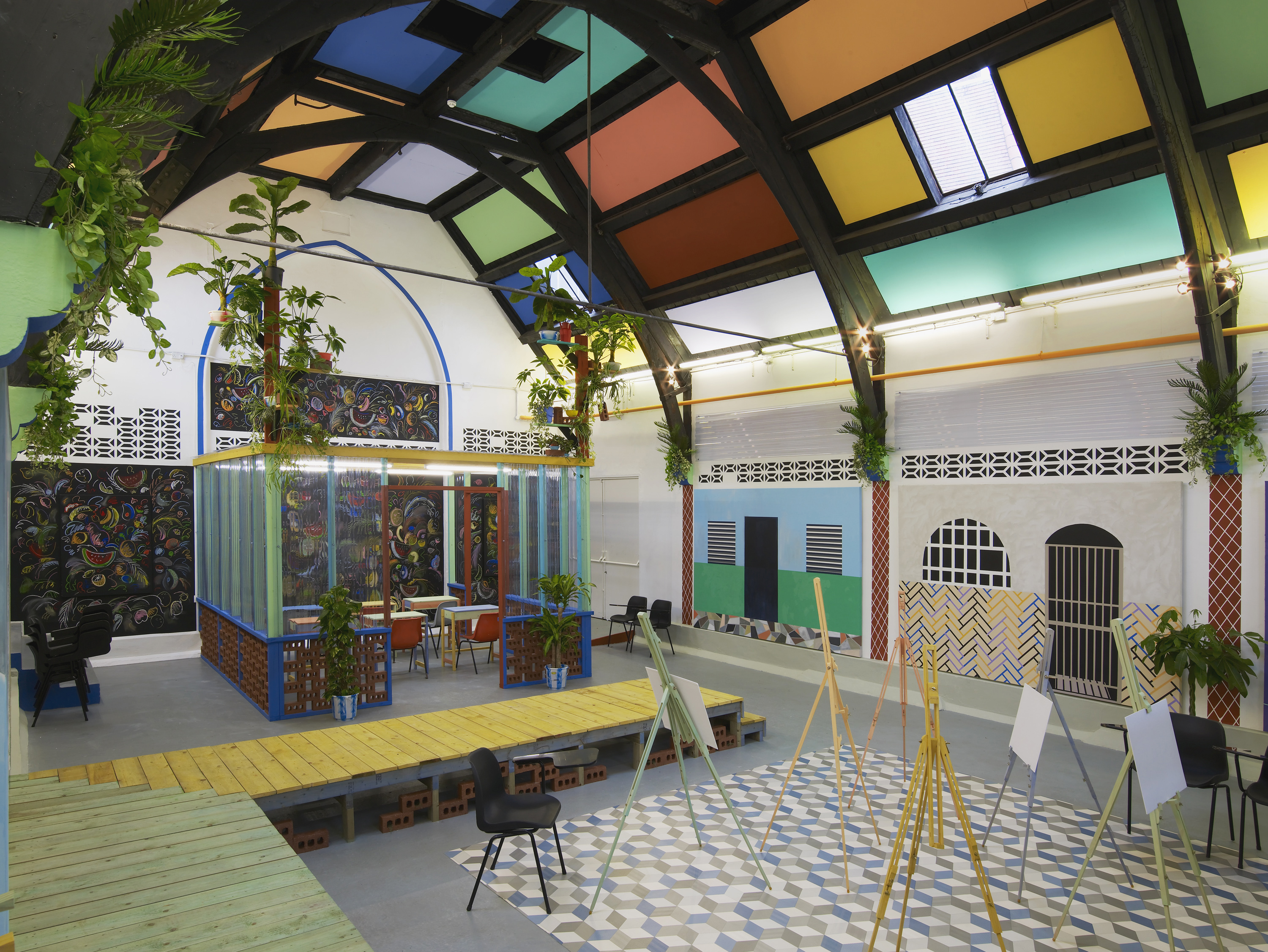 Installation view of Sol Calero’s exhibition at Studio Voltaire, showing a brightly coloured indoor art studio. The space features easels, potted plants, geometric floor patterns, and vibrant wall murals. An arched ceiling is segmented into multicoloured panels, creating a dynamic and energetic environment.