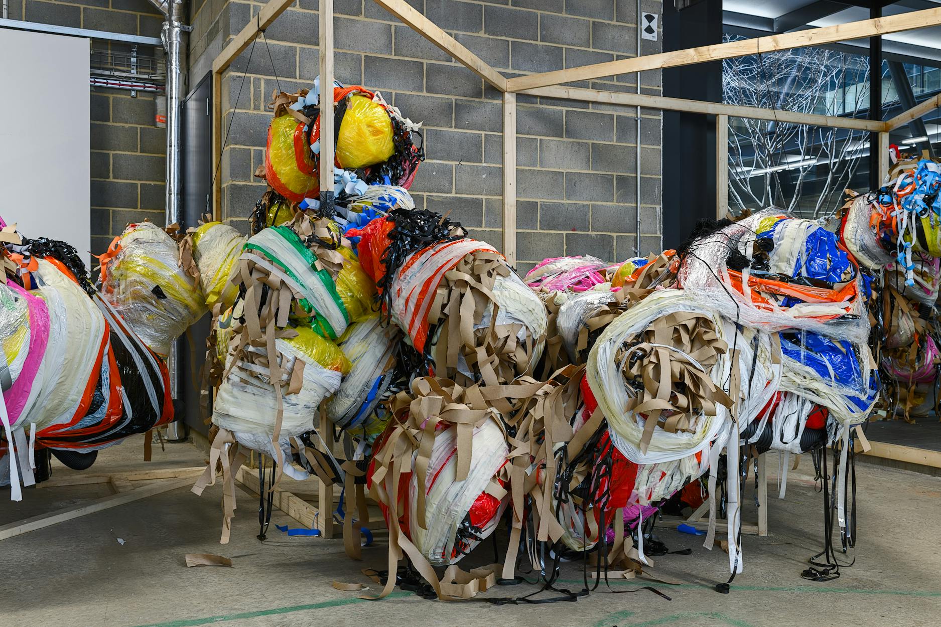 A sculpture made from bundles of colourful plastic sheets, fabric, and tan paper strips stands in an industrial space with concrete floors and breeze block walls, partially framed by an unfinished wooden structure.