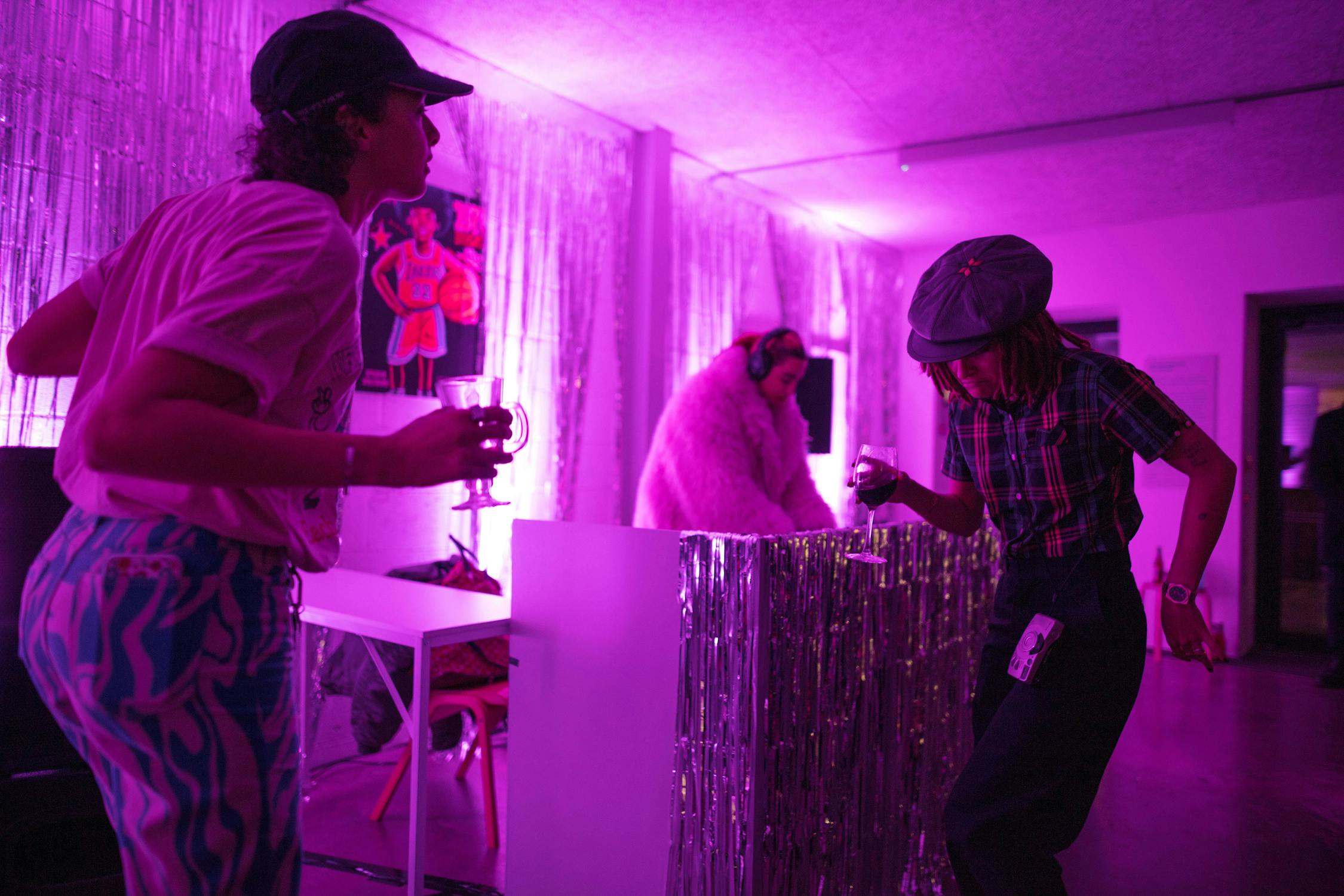 Two people dance in front of a woman dj, who wears a white fur coat. The room is lit with purple lights, and the walls are covered in silver tinsel.