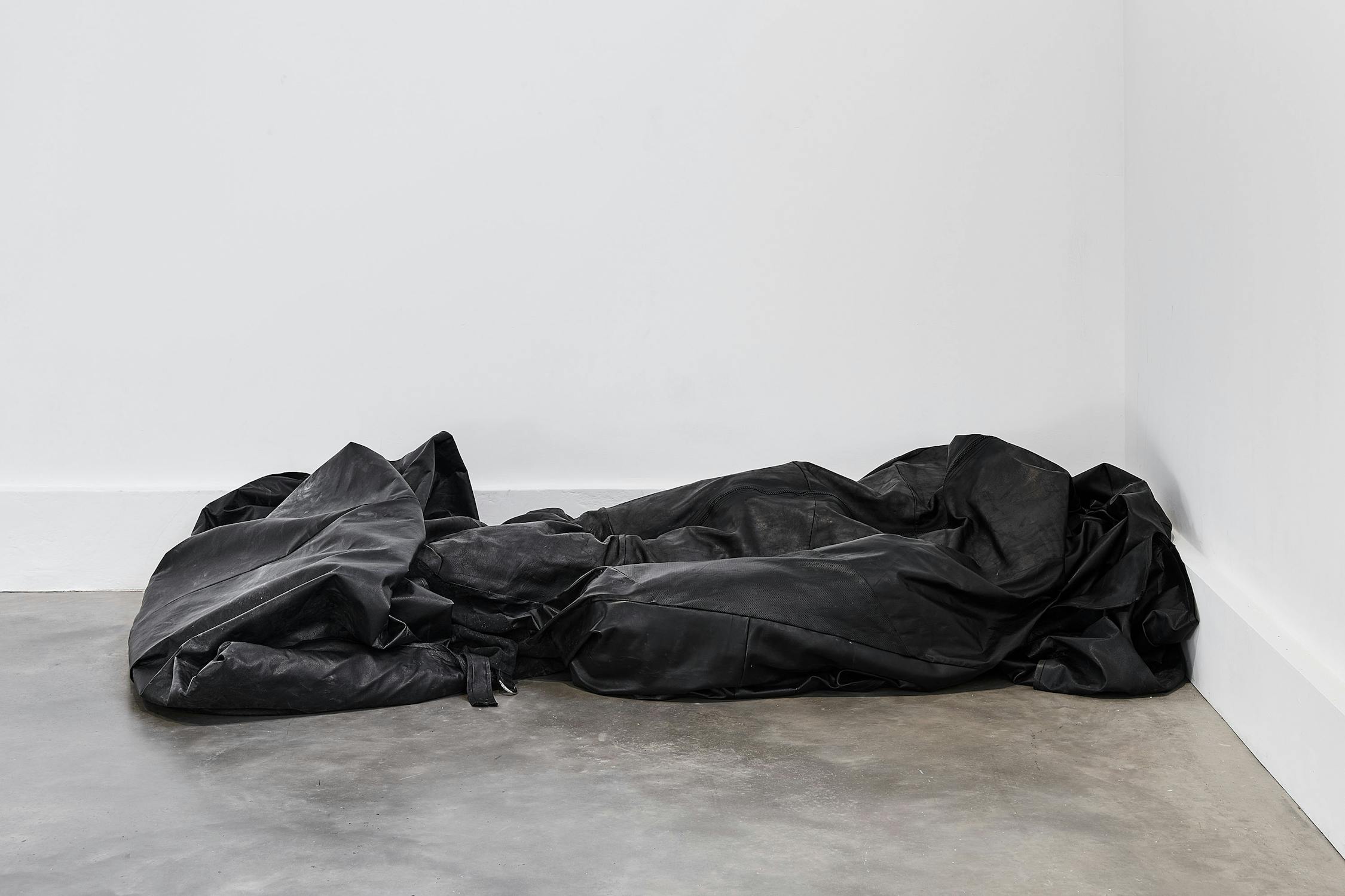A sculpture by Solomon Garçon. A large, crumpled black tarp or fabric lies on a bare concrete floor in the corner of a white-walled room, creating an abstract, sculptural form.