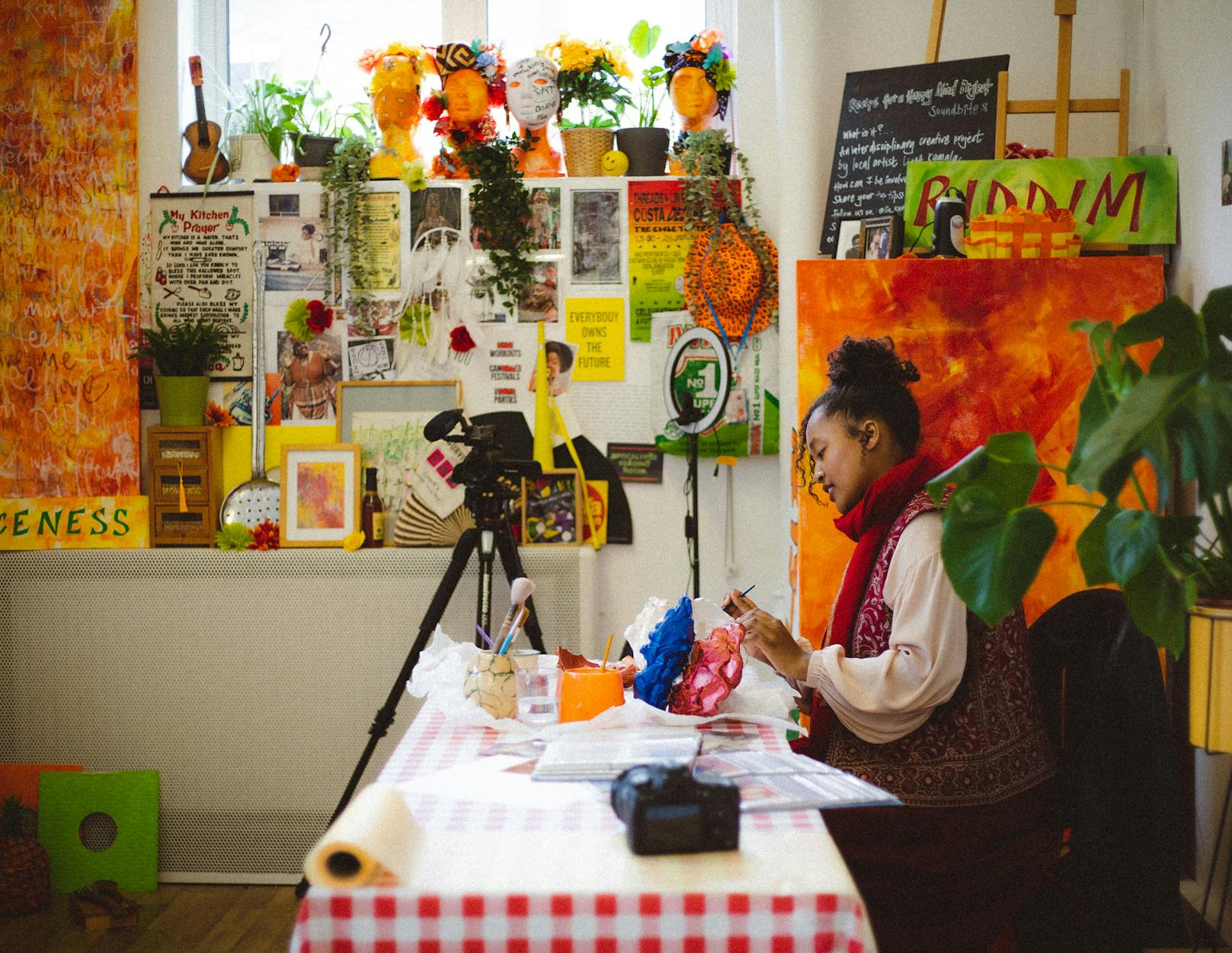 Artist Maya Gurung-Russell Campbell sits at a table covered with art supplies, crafting a colorful item. The room is vibrant, with various art pieces, plants, and bright decorations adorning the walls. A large canvas with orange tones stands in the background.