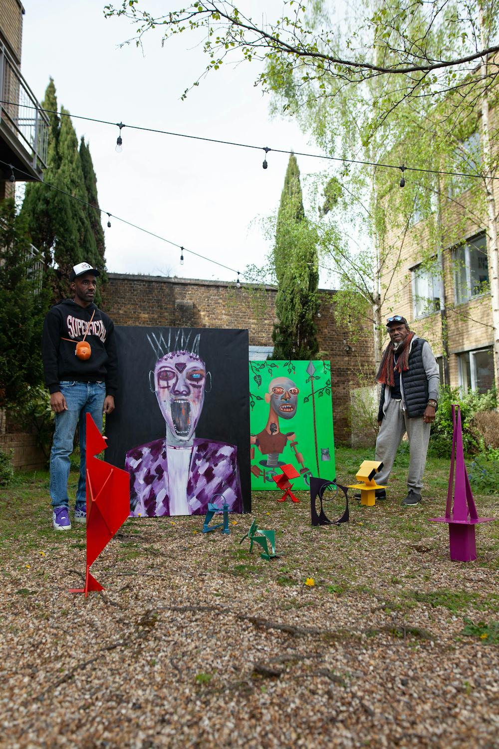 Two men stand in a courtyard with string lights, trees, and brick buildings. In front of them are two large colourful paintings—one of a stylised face and one of a figure—and several small abstract metal sculptures on the gravel ground.