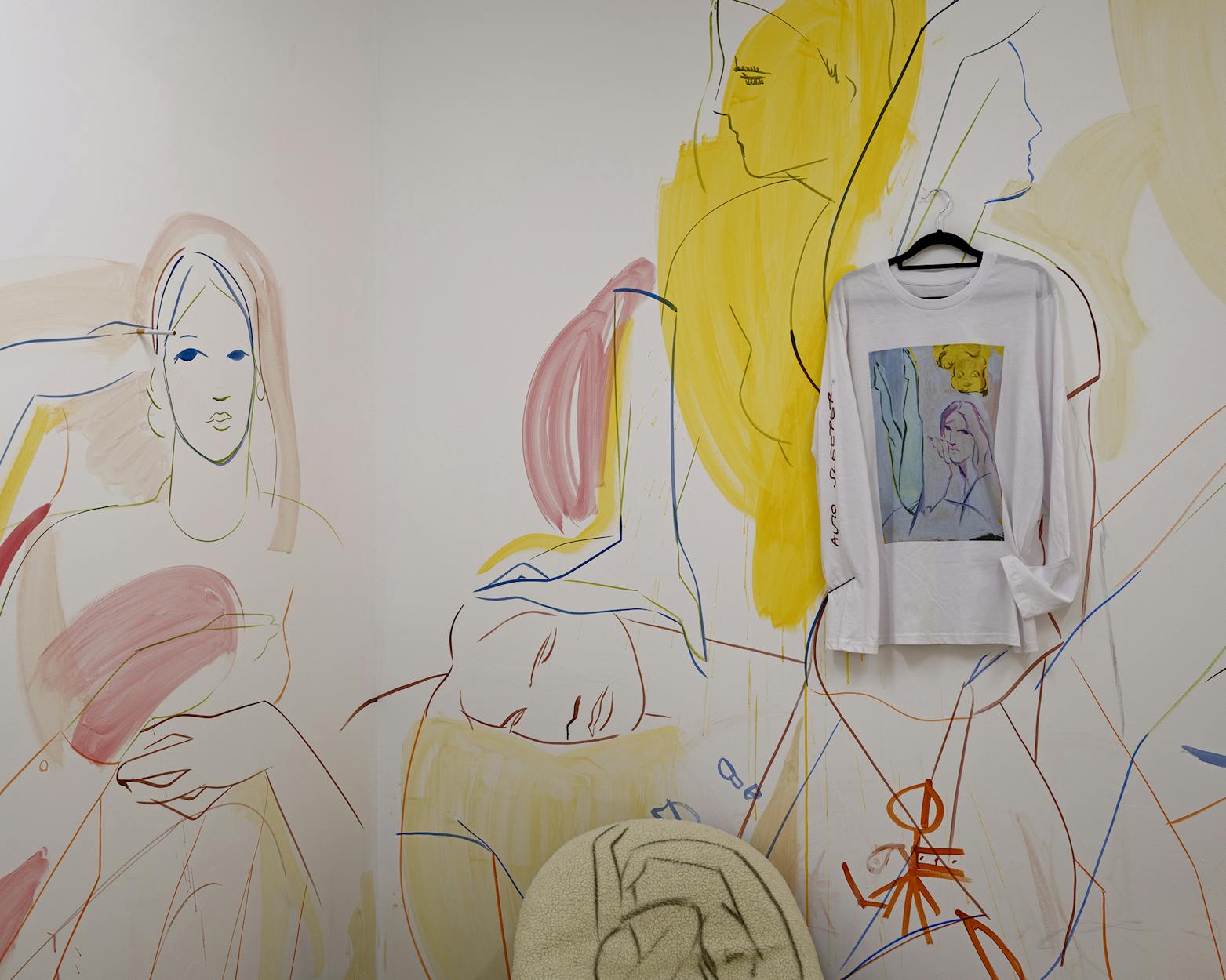 A wall features a colourful abstract mural of reclining figures. A white long-sleeve shirt with a similar illustrated design hangs on the wall. Below, a beige textured fabric is draped over a chair.