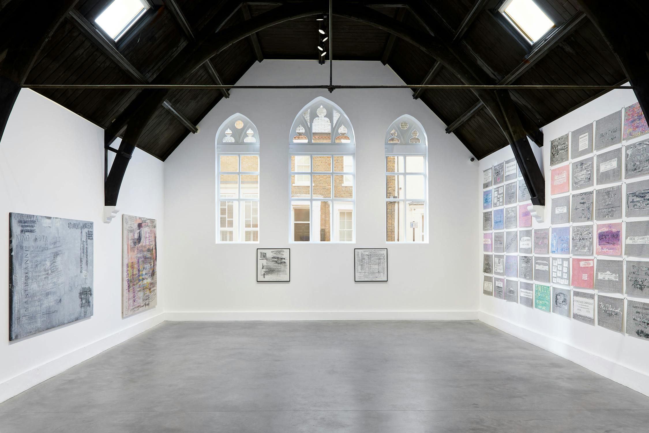 A gallery with white walls and a black vaulted ceiling. The walls are mounted with large chalf rubbings on canvas on the left and back wall. On the right wall there is a grid of smaller pencil drawings.