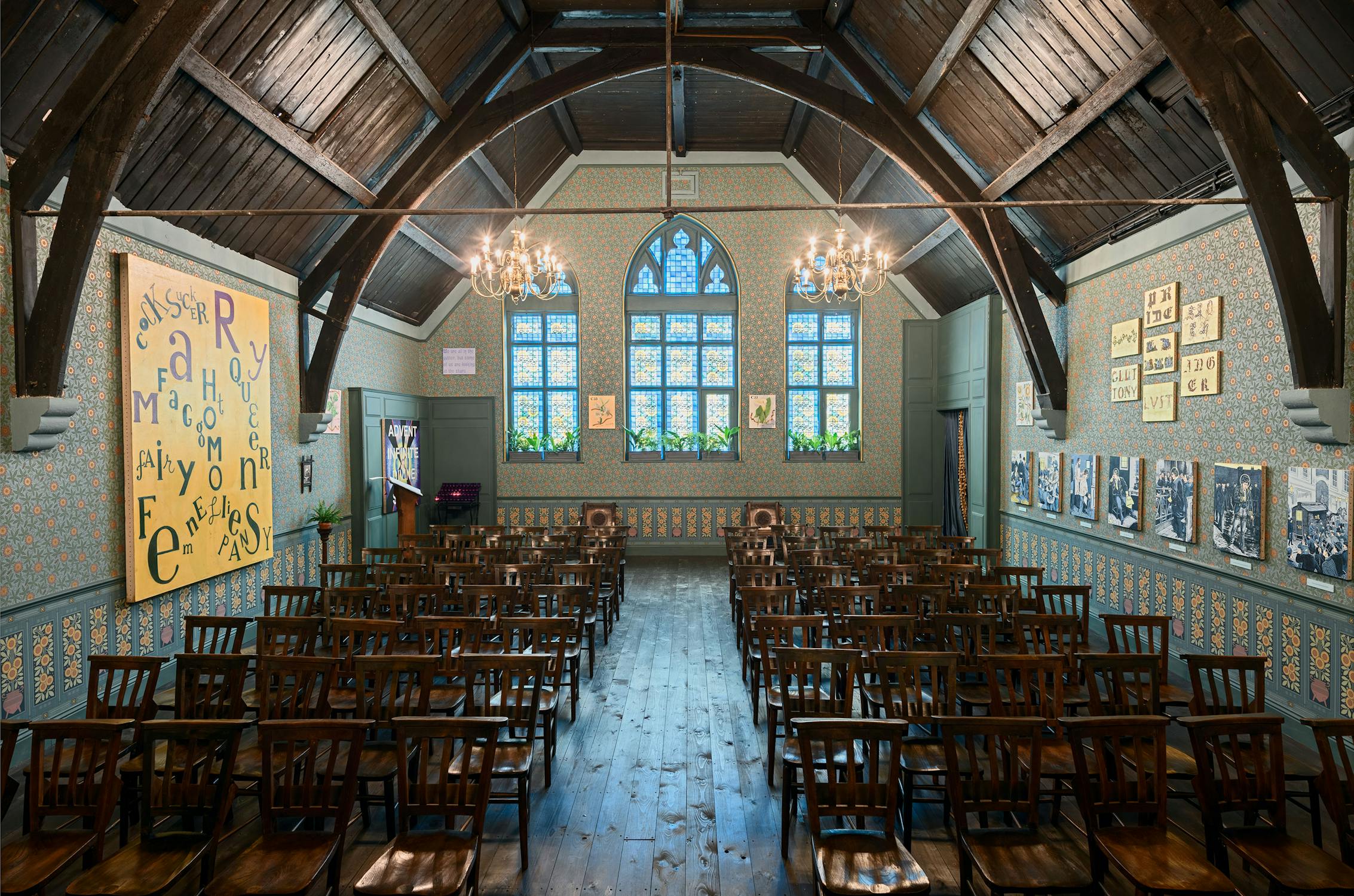 An historic room with wooden chairs facing a small stage adorned with a statue and floral arrangements. The walls are decorated with blue patterned wallpaper, chandeliers, framed art, and a large yellow board with jumbled letters in black and green.