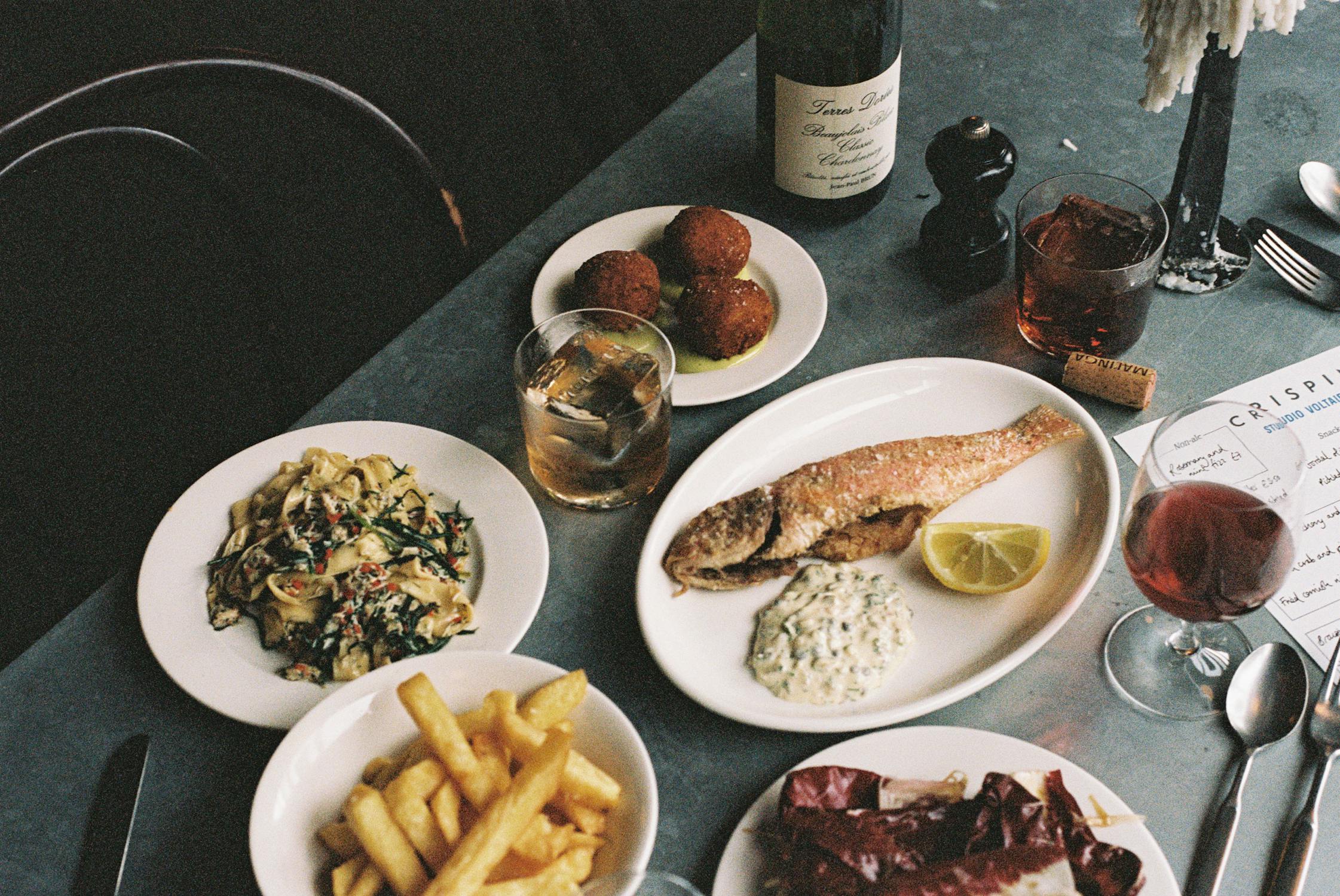 A photograph of a table with plates of food and drinks. There is a bowl of chips, a fried fish, croquettes, and different drinks that include red wine, a cocktail and a menu.