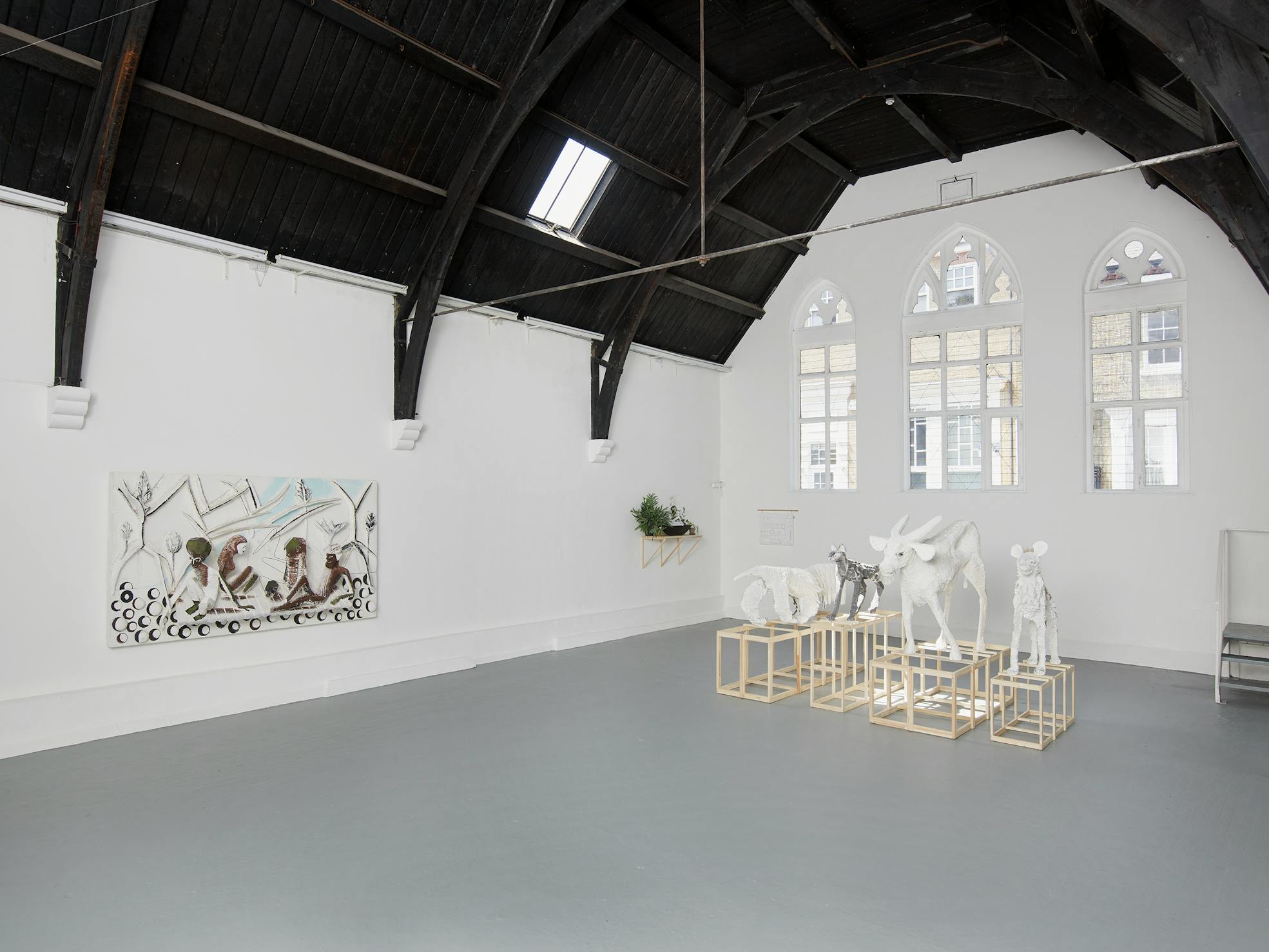 A bright art gallery with high arched windows and black beams displays four abstract animal sculptures on wooden cubes and a large textured relief artwork on a white wall. Light streams in, highlighting the modern, minimalist interior.