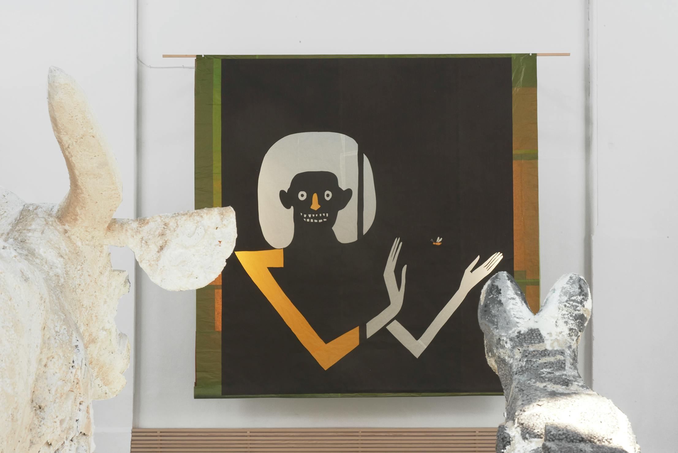 A black tapestry features a stylised figure with pale hair, a wide smile, and skeletal fingers, gesturing with arms bent. Two abstract pale sculptures, one cow-like and one cat-like, are in the foreground, partially blocking the view.