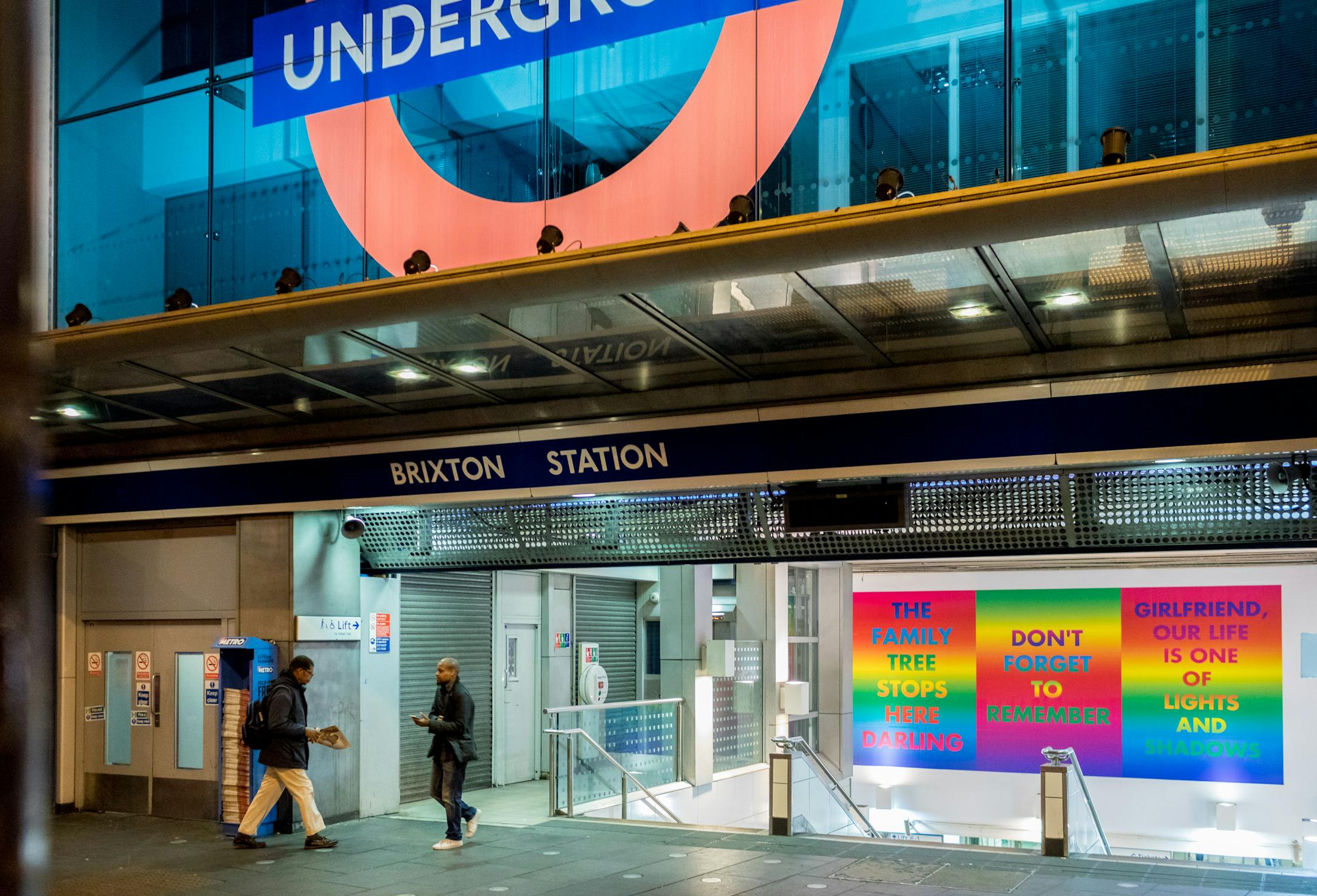 Two people walk past the entrance of Brixton Station at night. Above, a large London Underground sign is visible. Colourful posters with various positive messages are displayed within the station entrance.