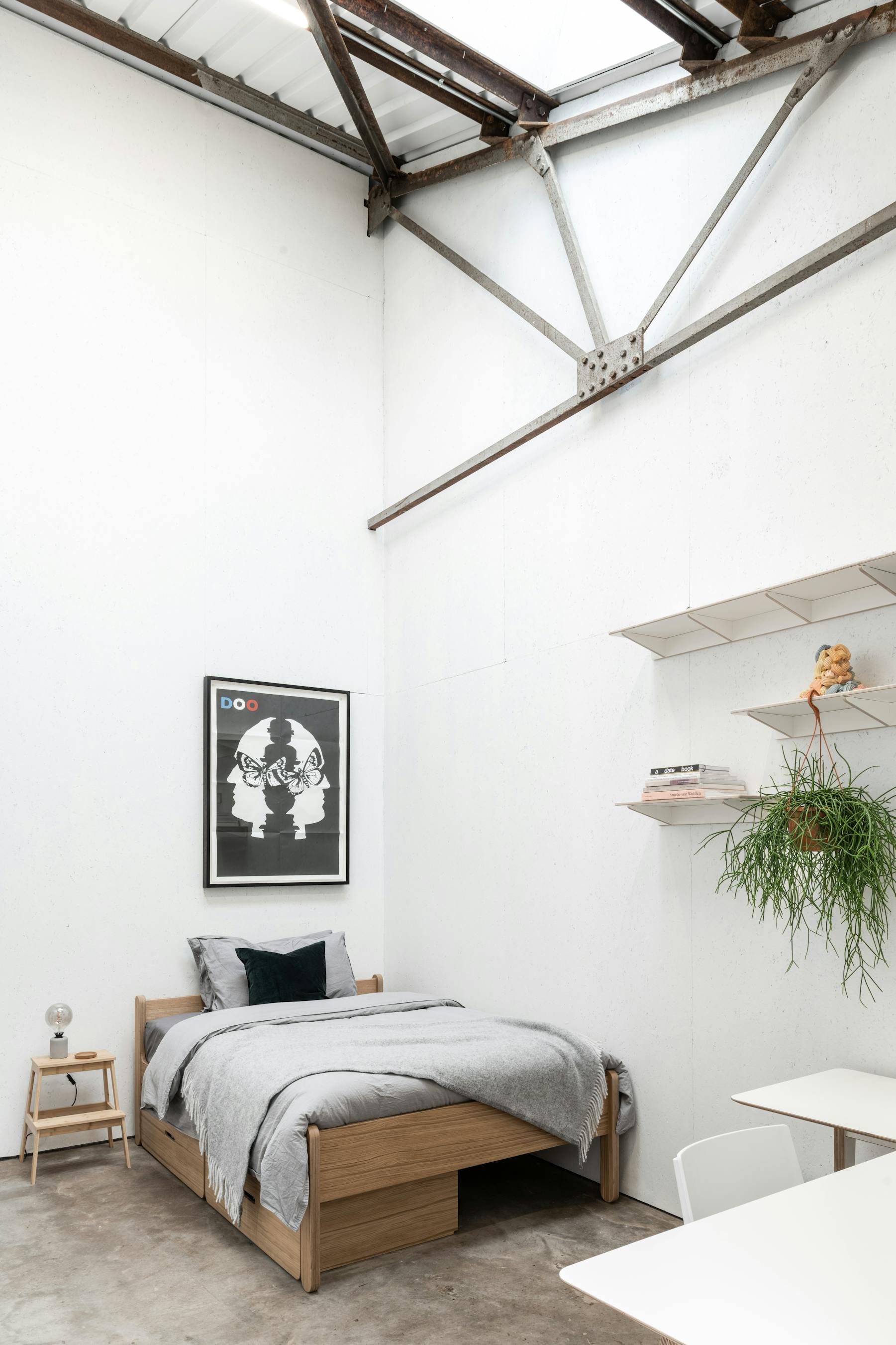 Minimalist room with white walls and high ceiling. There’s a single bed with a grey blanket, a corner desk with a white chair, a lamp, small potted plants, and shelves holding books, towels, and a plant. Floor is concrete, decor is simple and modern.