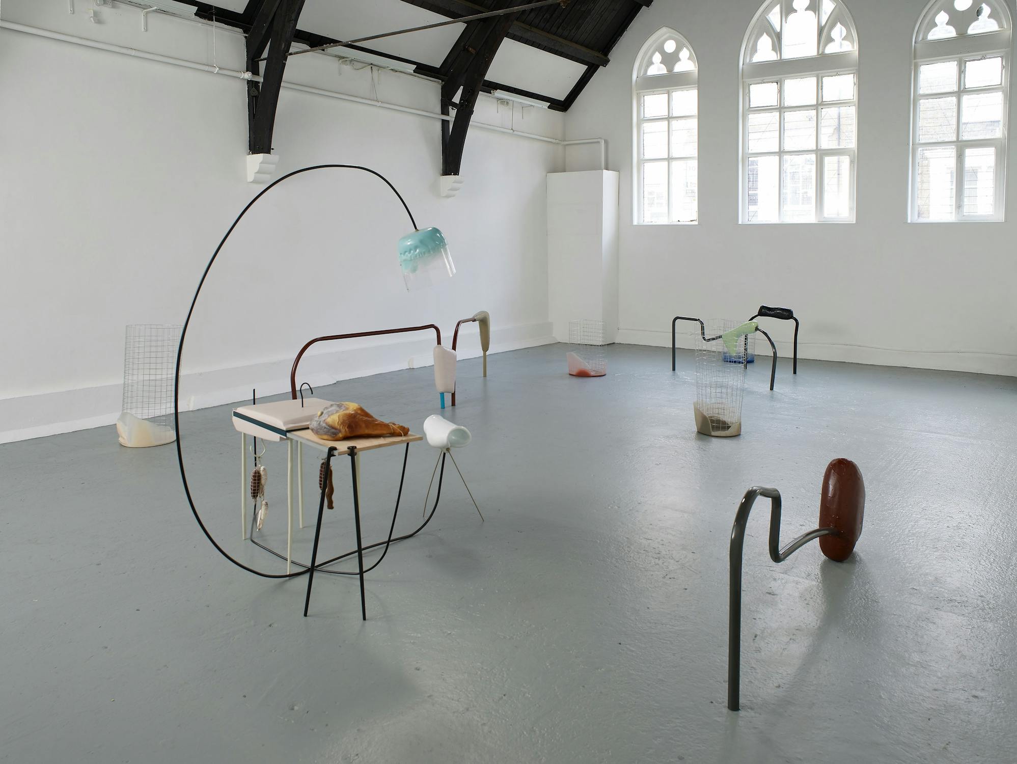 An art gallery with abstract sculptures, featuring metal rods, a table, a glass container, and fabric-like materials on a light grey floor. Gothic windows with arched tops let in natural light from the right side of the room.