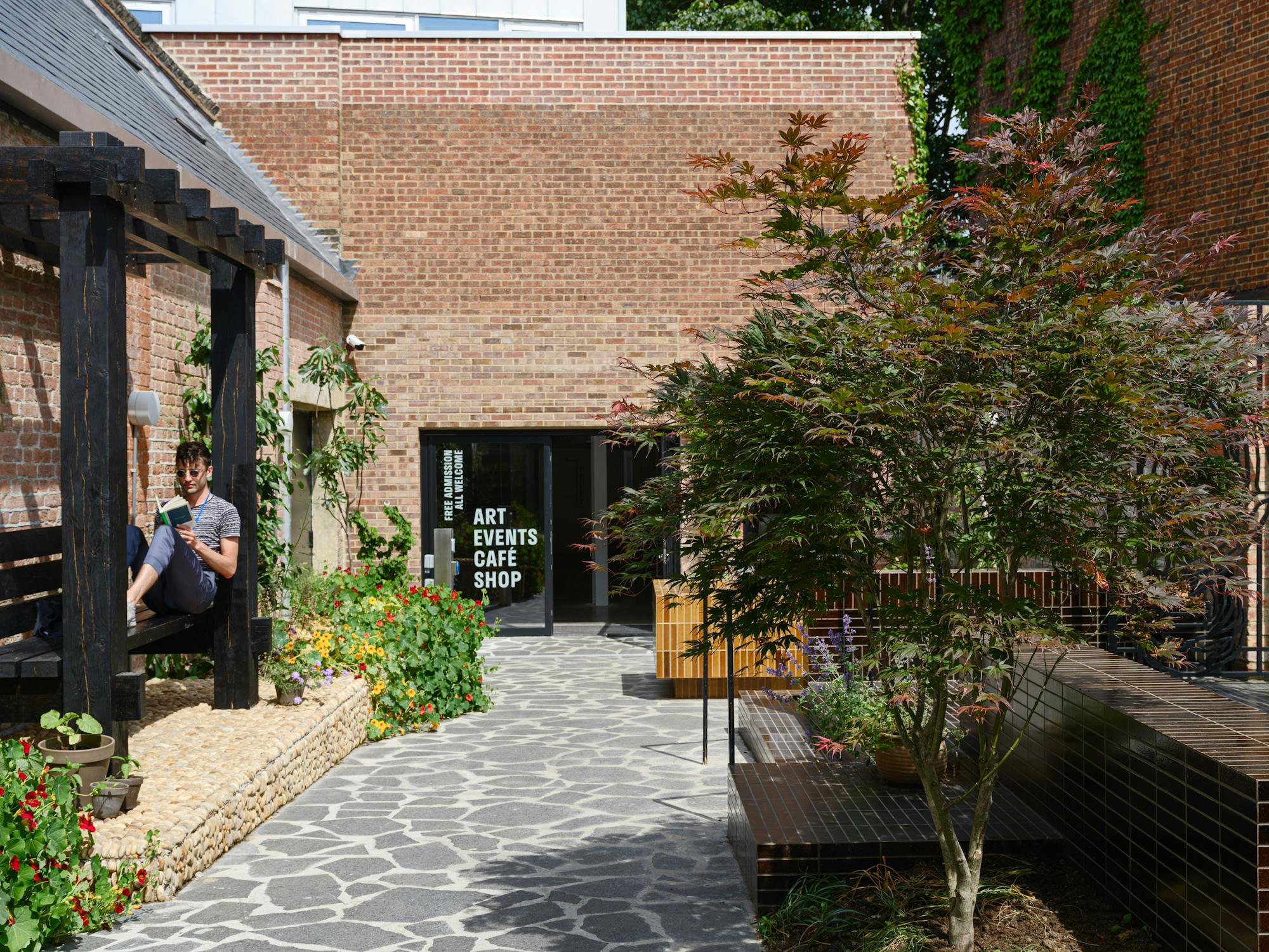 Studio Voltaire's garden in summer, photographed on a sunny day. The garden is paved with irregular slabs that slope towards the entrance. a man sits on a black wooden pergola, reading a book.