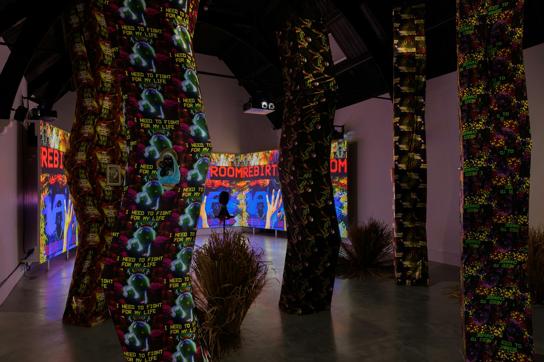A photograph of a dimly lit room, that is filled with tall angular structures that reach from the floor to the ceiling. The structures are wrapped in vibrantly patterned fabrics. In the bcakground, through the structure, viewers can see a large screen with colourful projections.