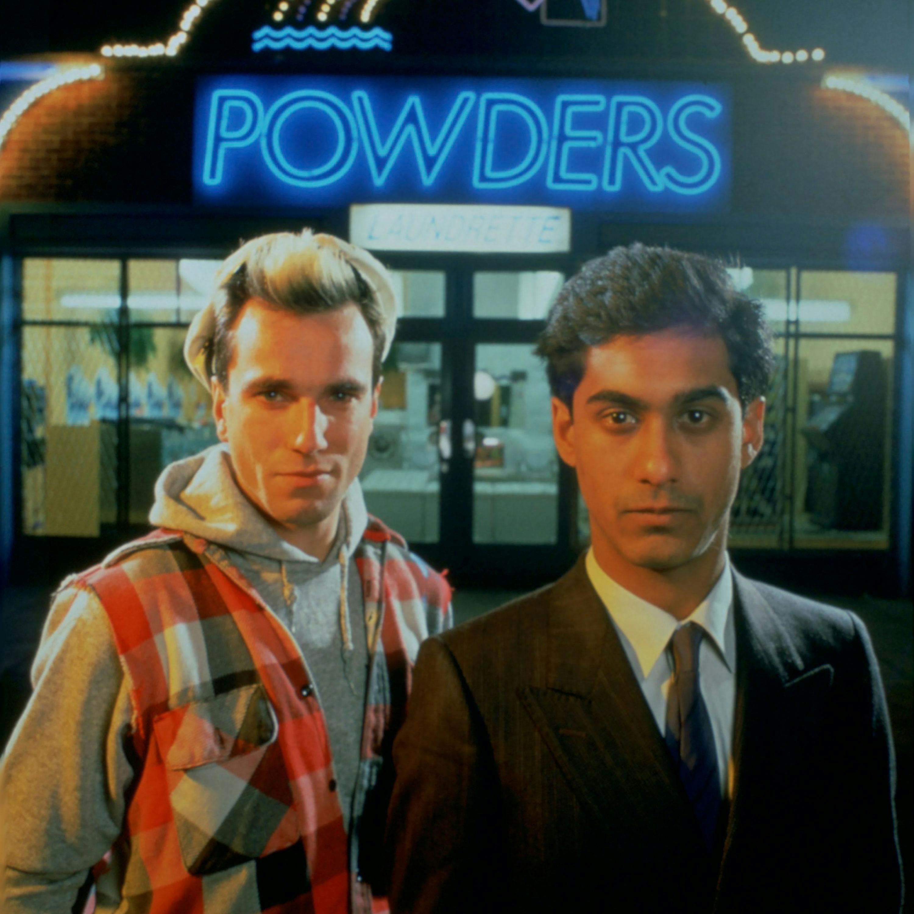 A white man wearing a grey hoodie and a flannel overshirt stands beside a south asian man in a brown suit and tie. they stand in front of. building with a neon sign that reads 'POWDERS'