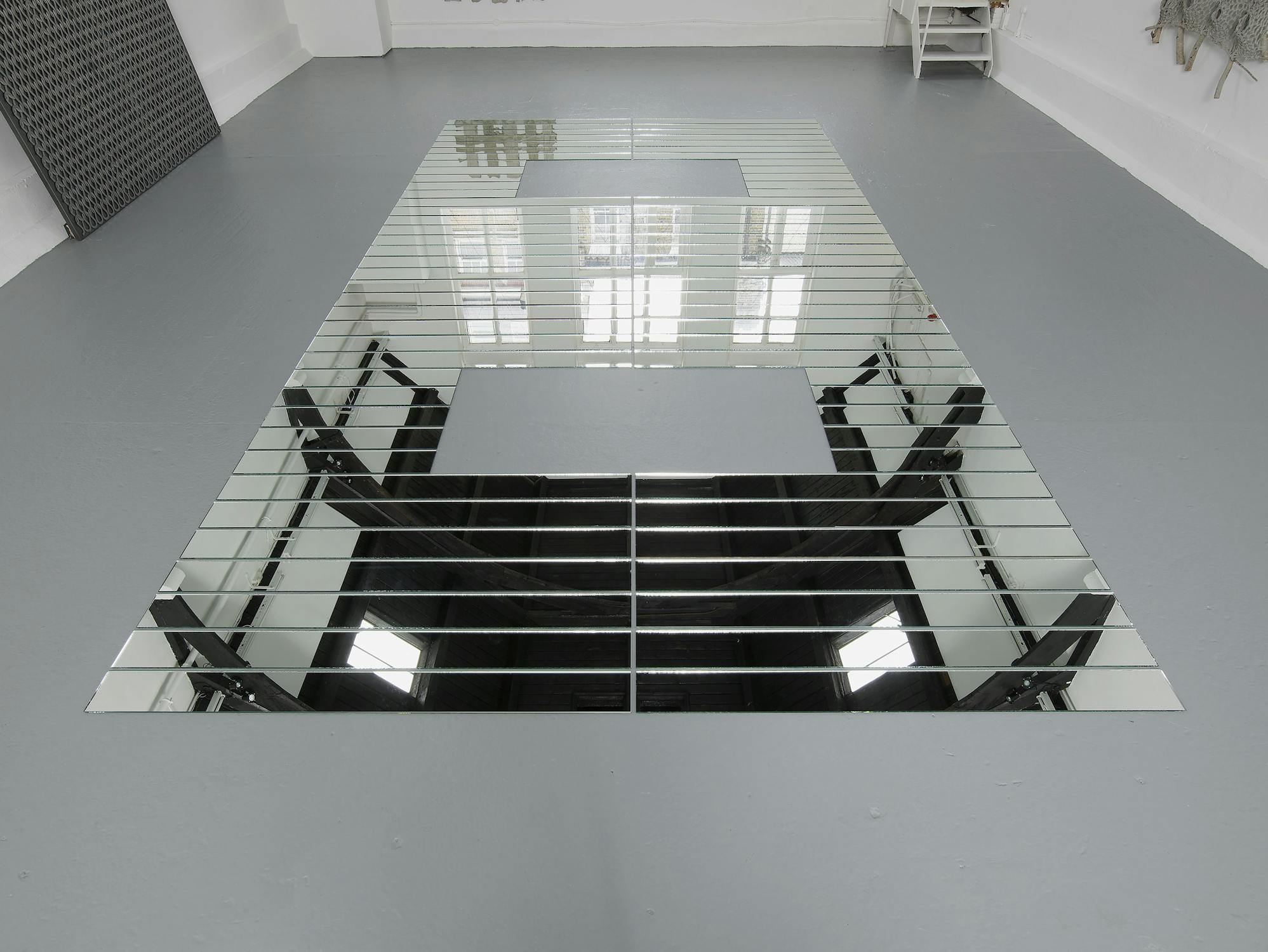 A geometric art installation on a grey floor features rectangular mirrors arranged in a large, hollow rectangle with a smaller central opening. The mirrors reflect the white walls, windows, and ceiling lights of the minimal gallery space.