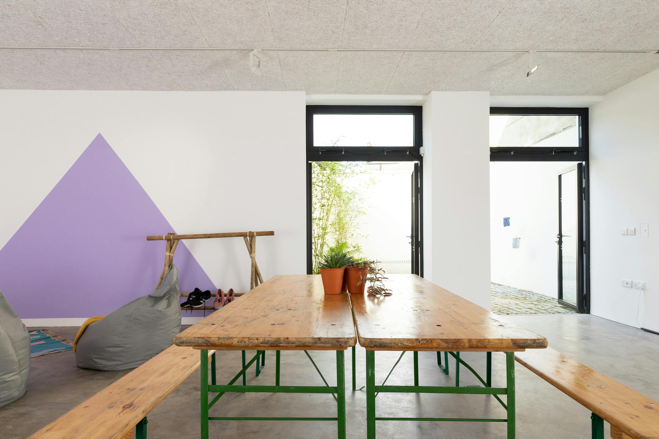 A bright room with a wooden table and benches, potted plants, bean bag chairs and a purple triangle painted on the wall. Two doors let in natural light and show greenery outside.
