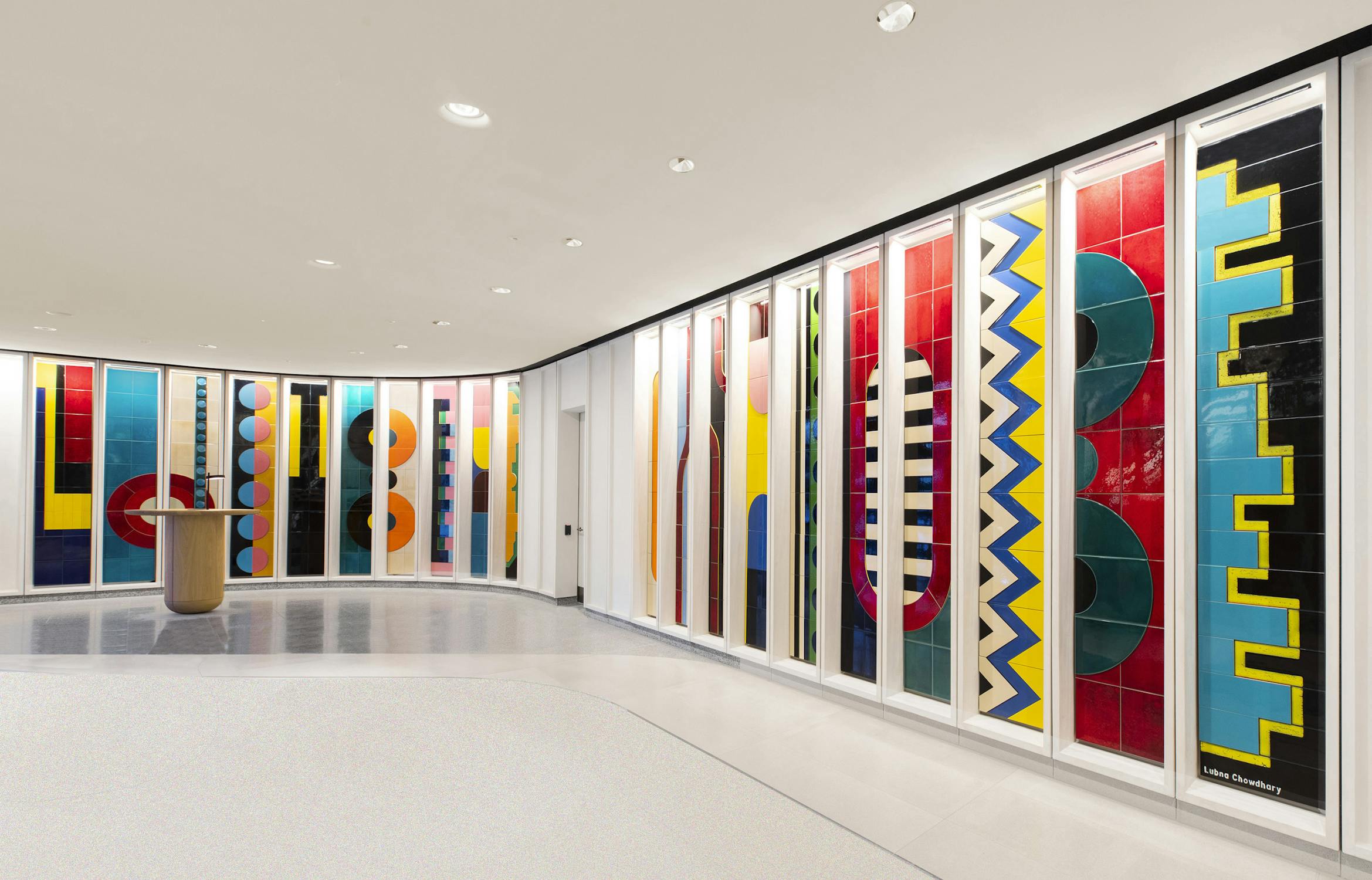 A colorful series of abstract murals on a curved wall, featuring geometric patterns in vibrant blue, red, yellow, and green. A tall, cylindrical wooden stand is placed nearby, with a smooth white floor and ceiling lights above.