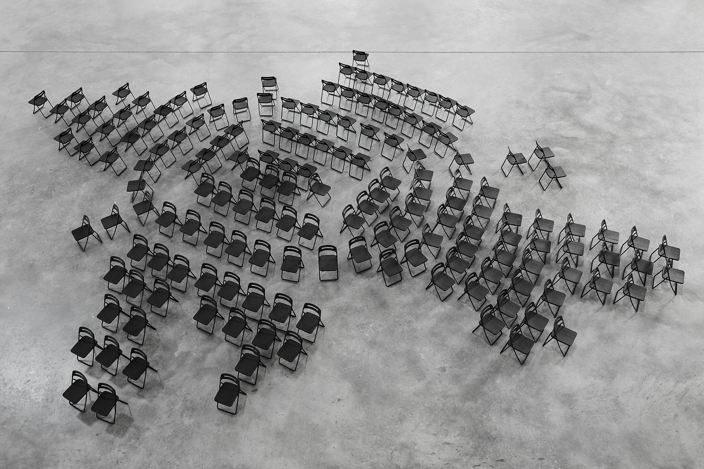 Rows of small 3D printed black folding chairs arranged on a large empty concrete floor, forming an abstract shape when viewed from above.