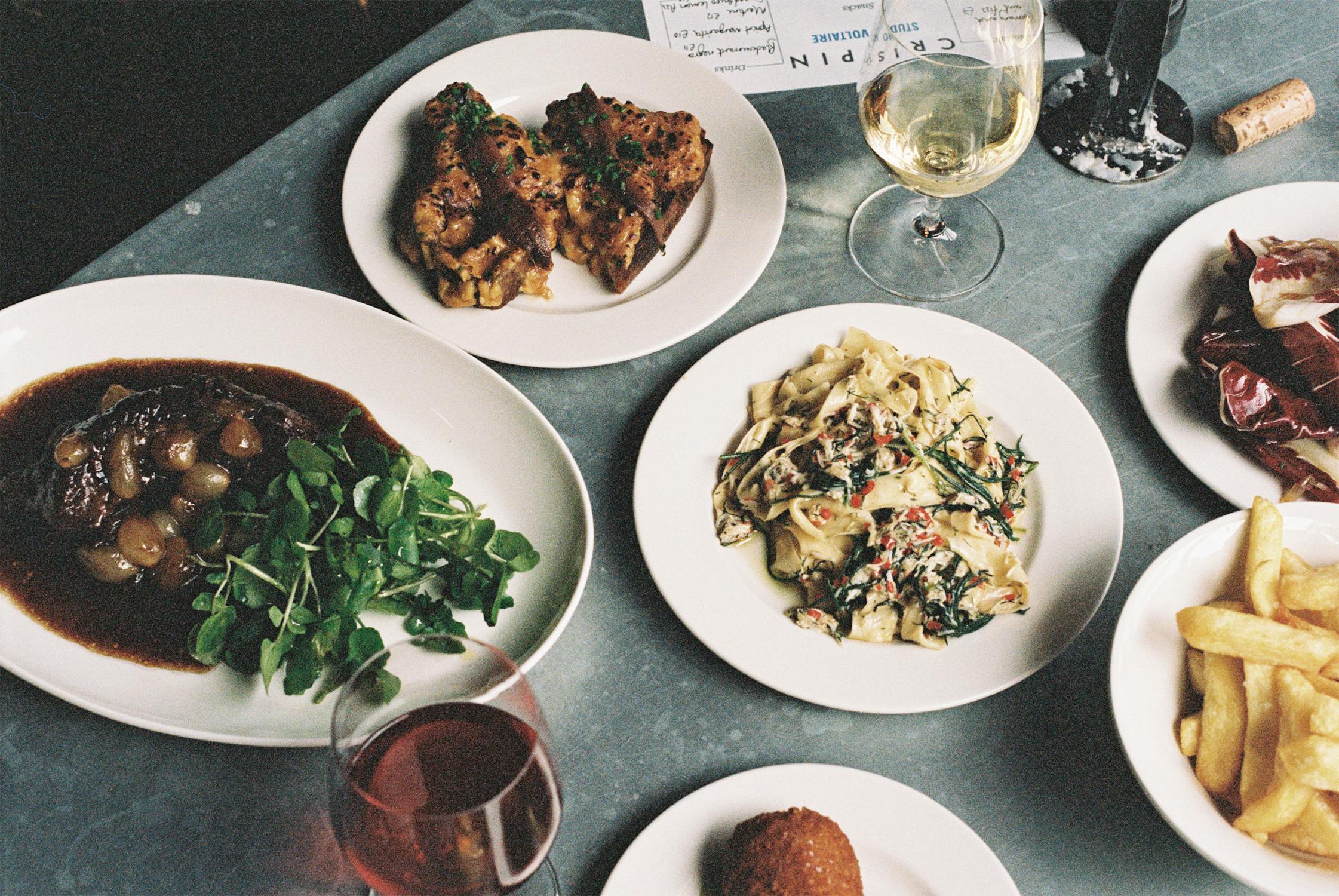 A photograph of a spread of freshly prepared restaurant food on white plates, including a creamy pasta dish, a joint of meat in gravy, a bowl of chips and glasses of red and white wine.