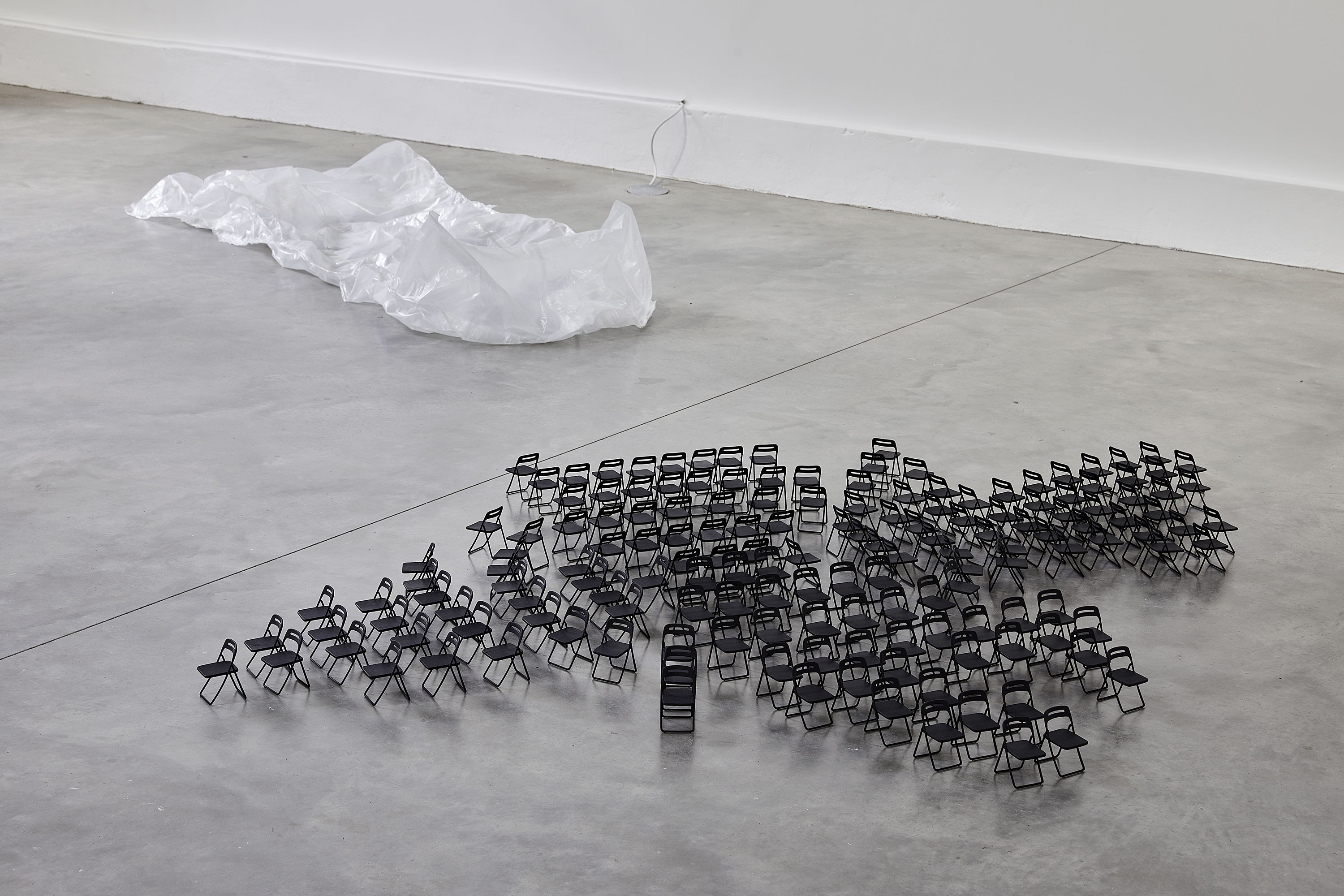 A group of small black folding chairs is arranged closely together on a grey concrete floor, with a large, crumpled sheet of transparent plastic lying in the background near a white wall.