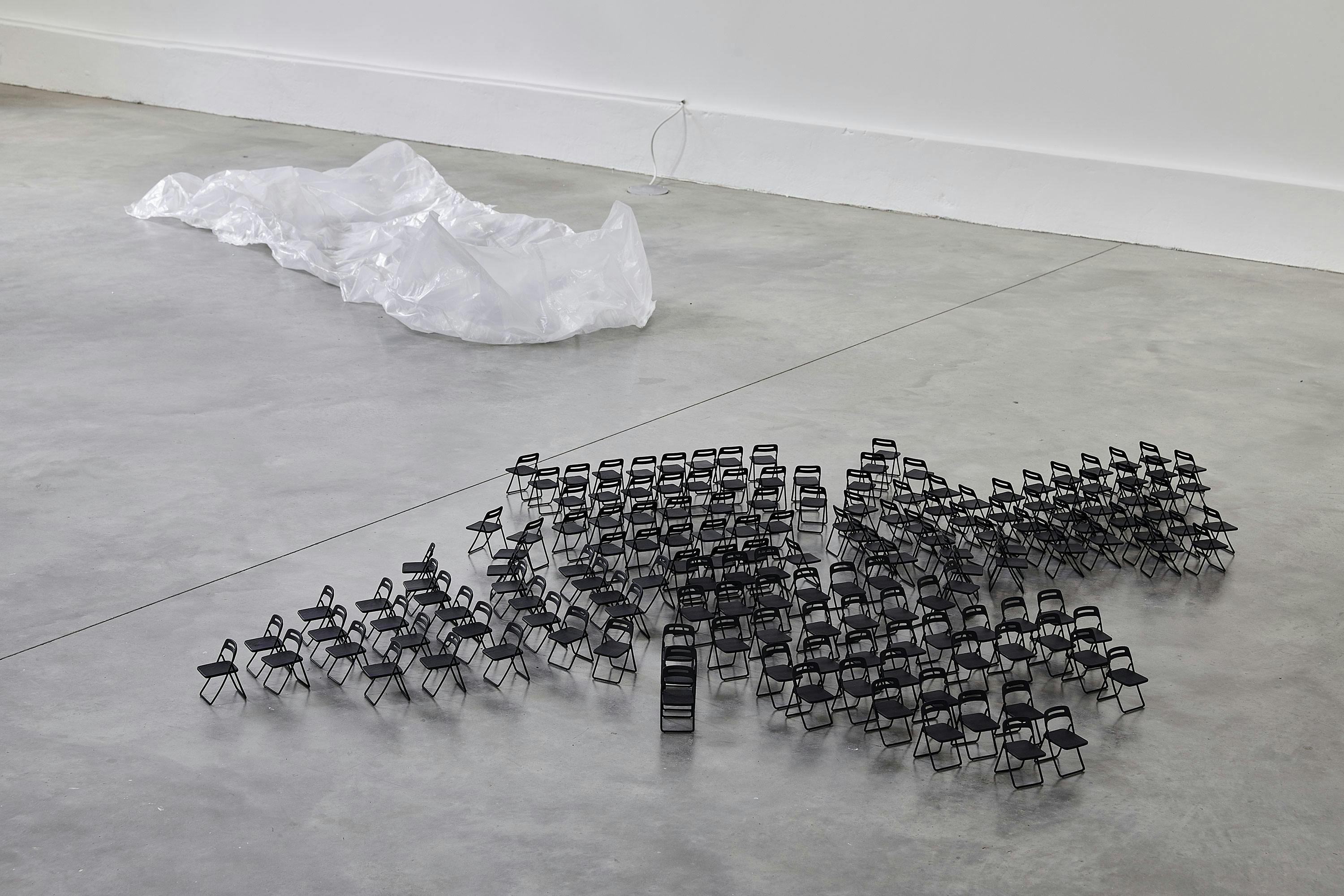 A group of small black folding chairs is arranged closely together on a grey concrete floor, with a large, crumpled sheet of transparent plastic lying in the background near a white wall.