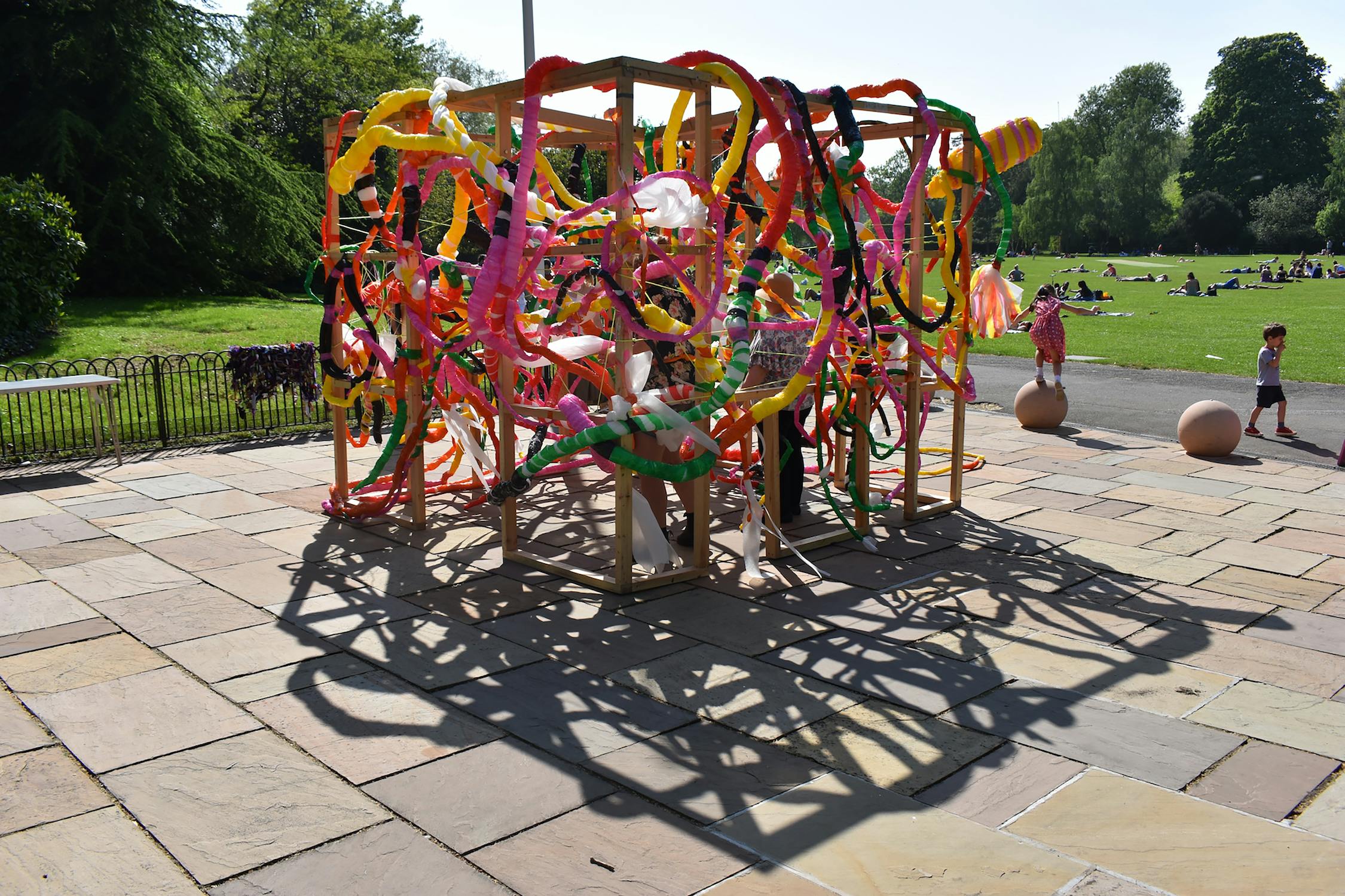 A colorful art installation made of intertwined, multicolored fabric strips forms a cube-like structure in a park. People interact with the piece, surrounded by green lawns and trees under a sunny sky.