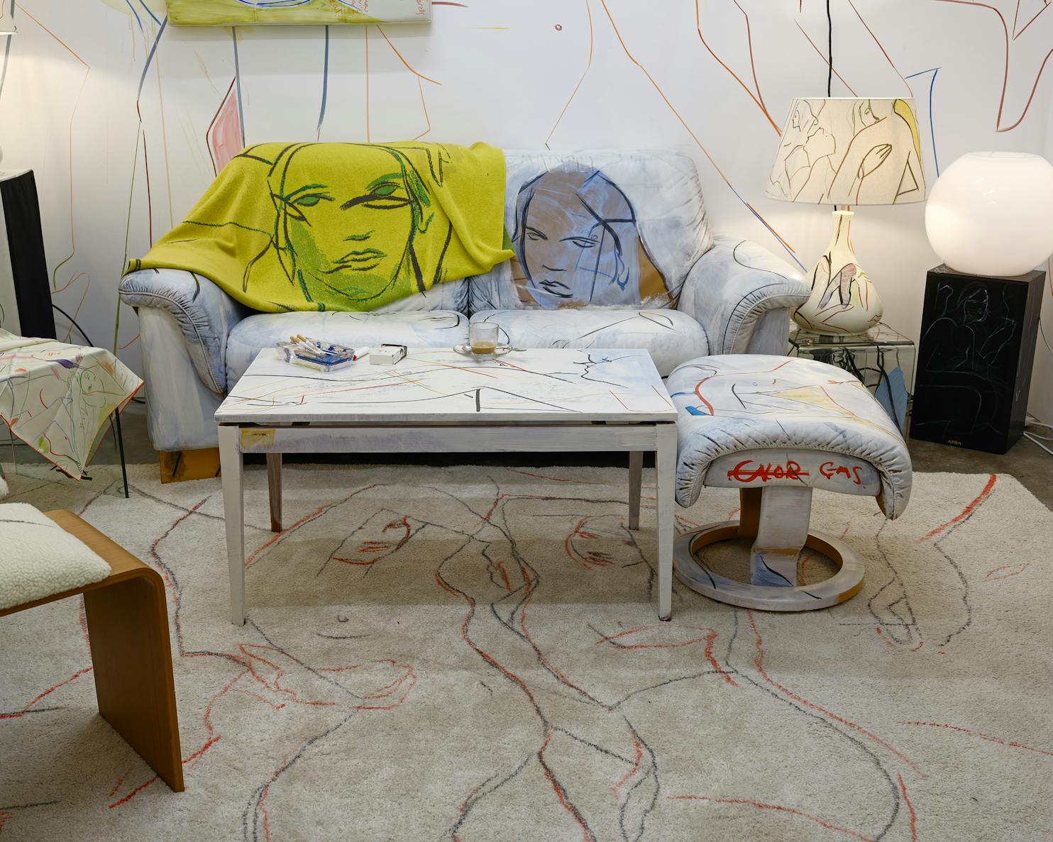 A living room featuring a white sofa draped with a yellow blanket, both printed with abstract line drawings of faces. A matching ottoman, table, and rug all display similar line art. Modern décor and soft lighting complete the scene.