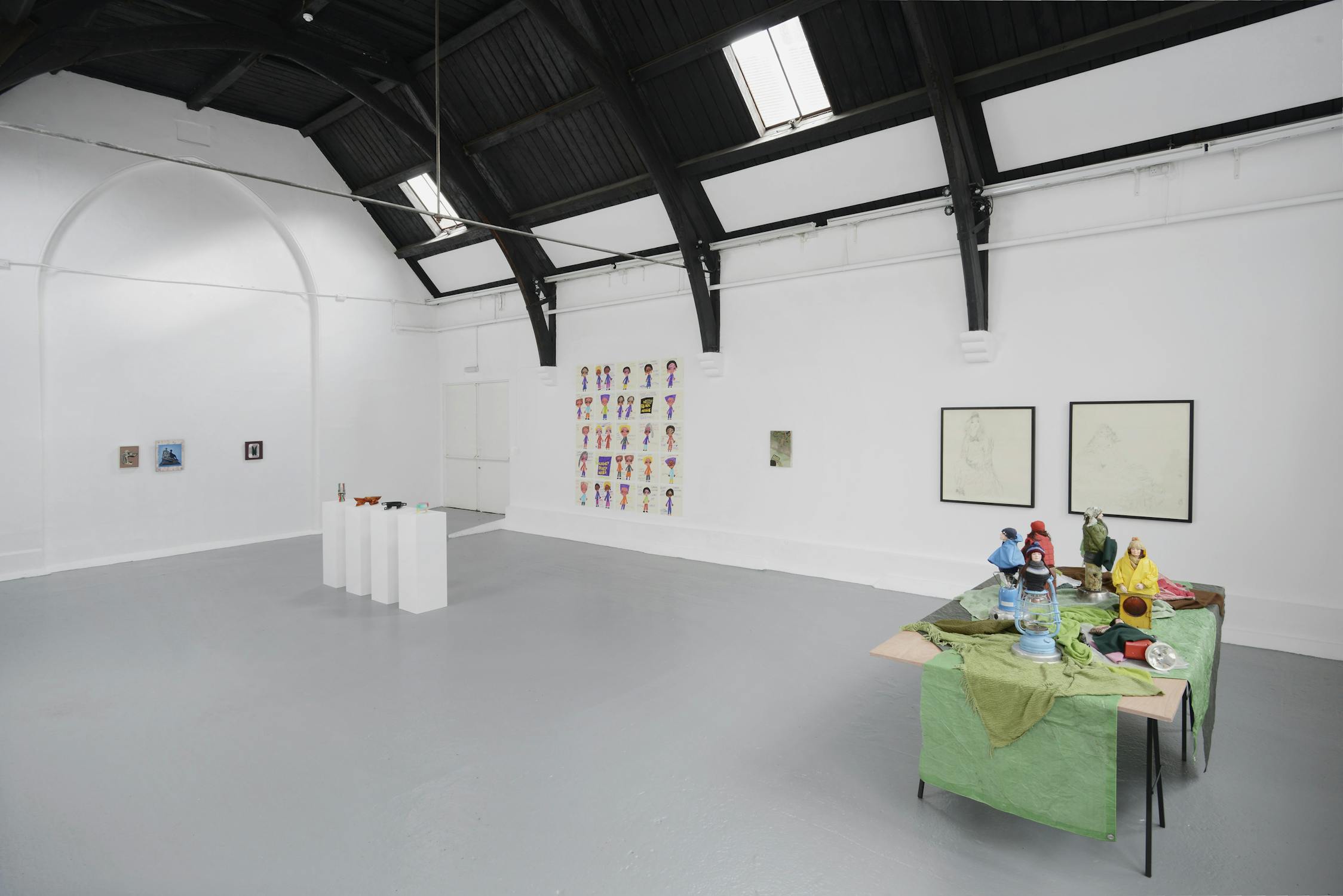 A contemporary art gallery with white walls and high, dark rafters features paintings, drawings, and colorful sculptures displayed on pedestals and a table covered with green fabric. Natural light enters through skylights.