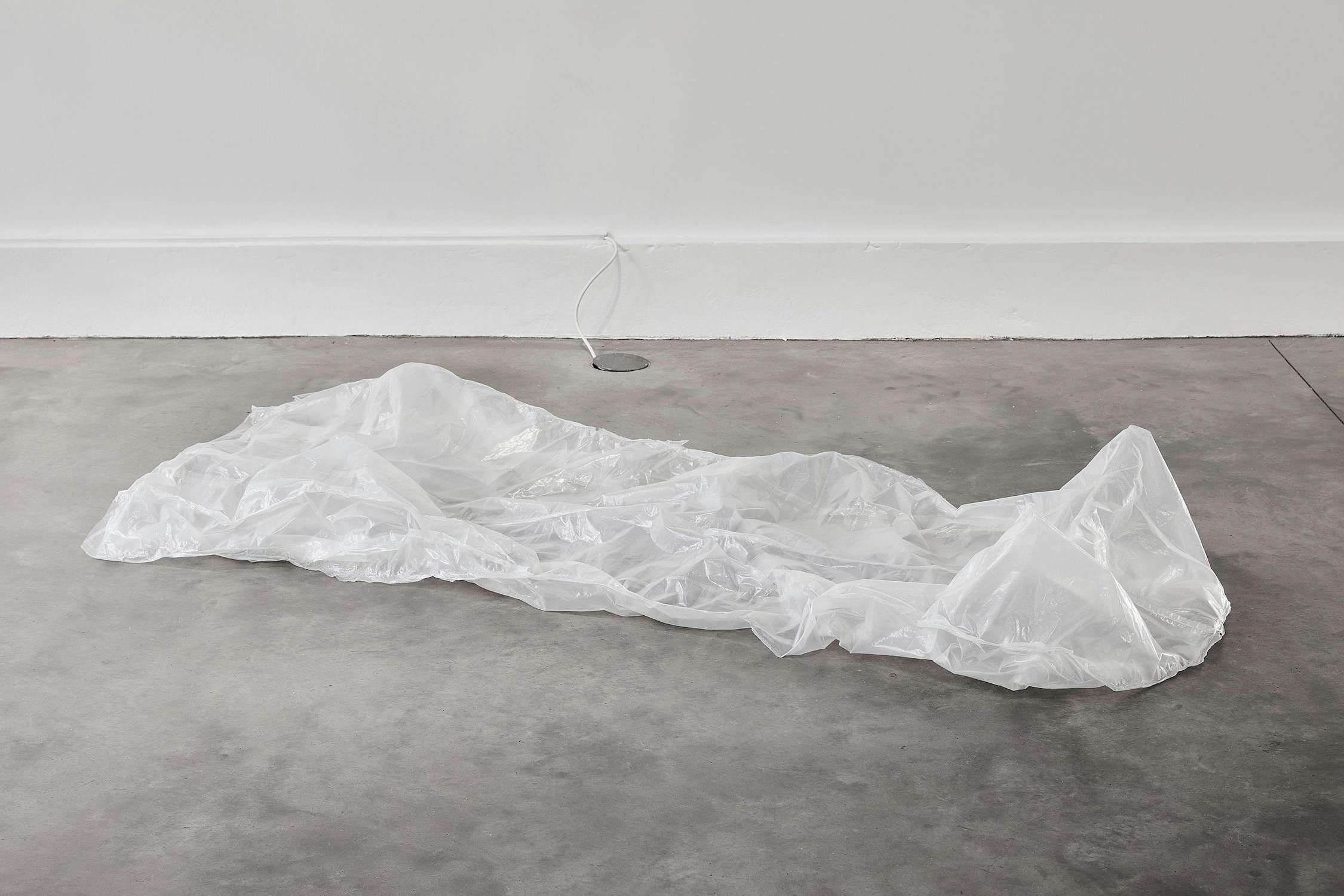 A crumpled, transparent plastic sheet lies on a grey concrete floor near a white wall with an electrical cord plugged into a socket in the background.