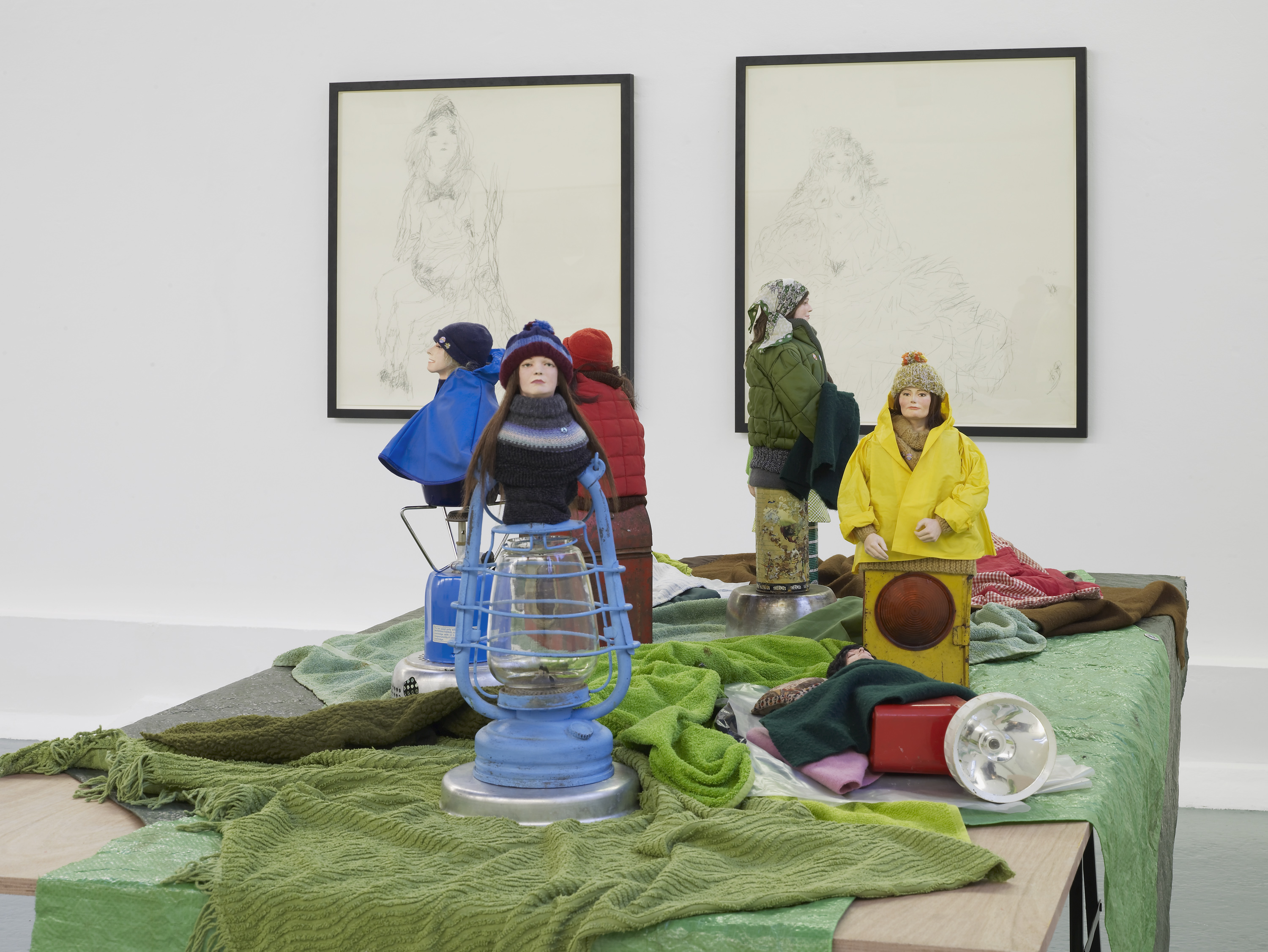 A group of small, lifelike dolls dressed in winter clothes are arranged on green fabric with various objects. Two framed sketches hang on a white wall in the background.