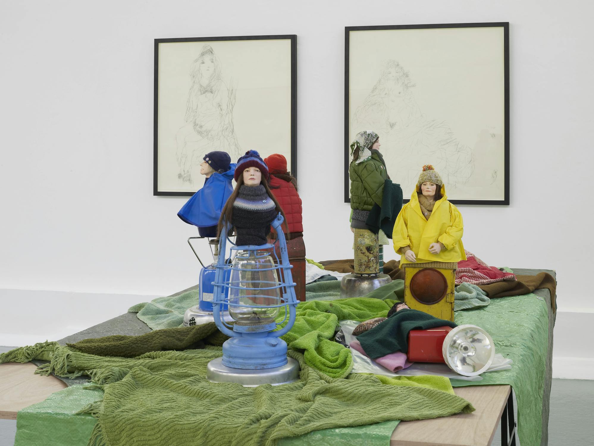 A group of small, lifelike dolls dressed in winter clothes are arranged on green fabric with various objects. Two framed sketches hang on a white wall in the background.