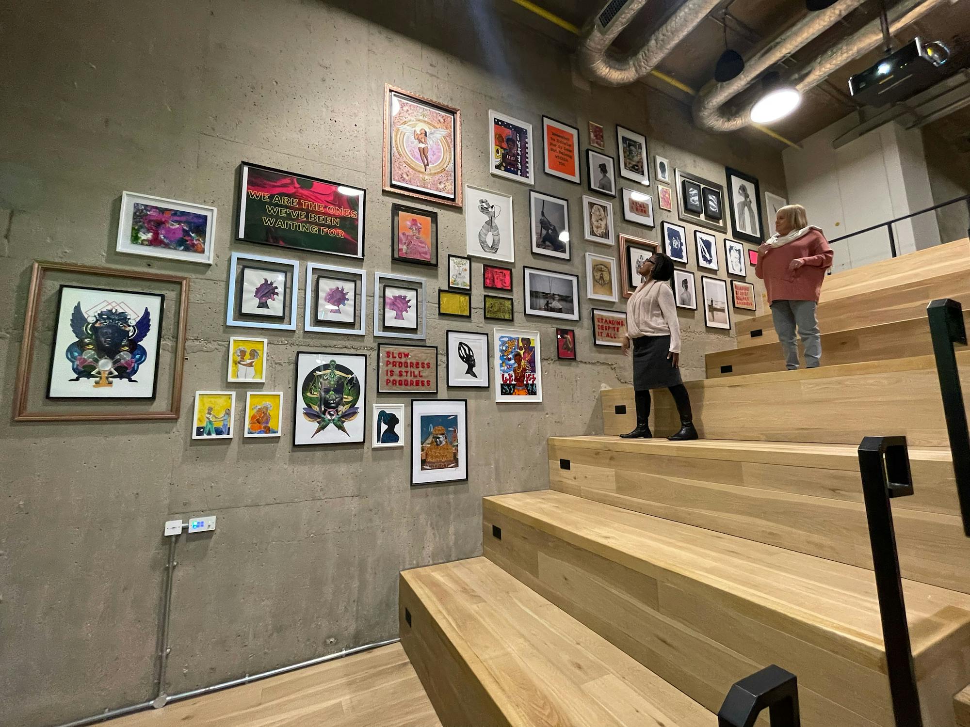 Two children walk up wide wooden steps with black railings in a modern, industrial-style space. A concrete wall to the left is decorated with an eclectic gallery of colourful framed artwork of various sizes and styles. Exposed pipes run along the ceiling.