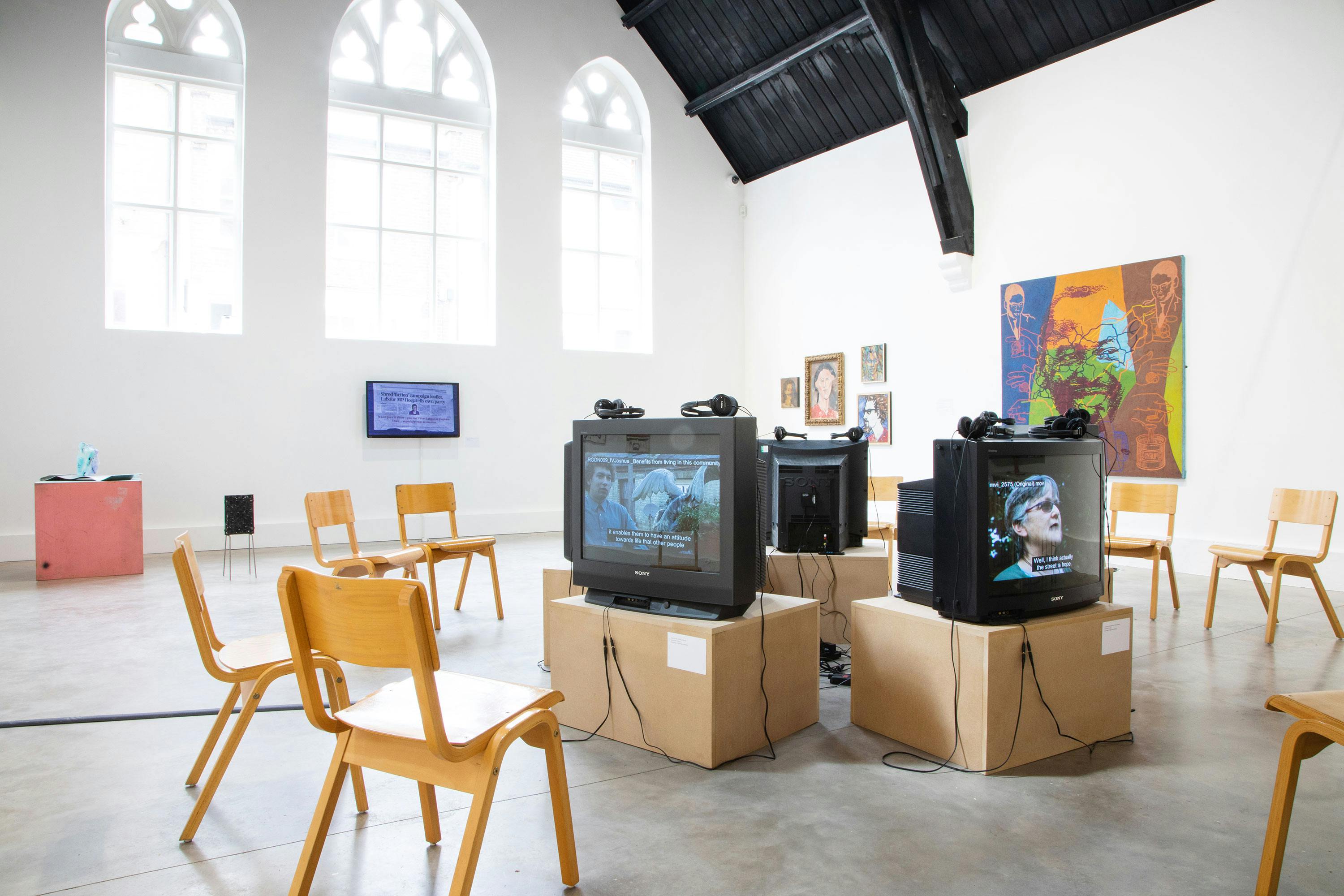 A modern art gallery with tall arched windows and black ceiling. Wooden chairs are arranged around vintage TVs placed on boxes. Various artworks, including paintings and small sculptures, are displayed on the walls and a pedestal.