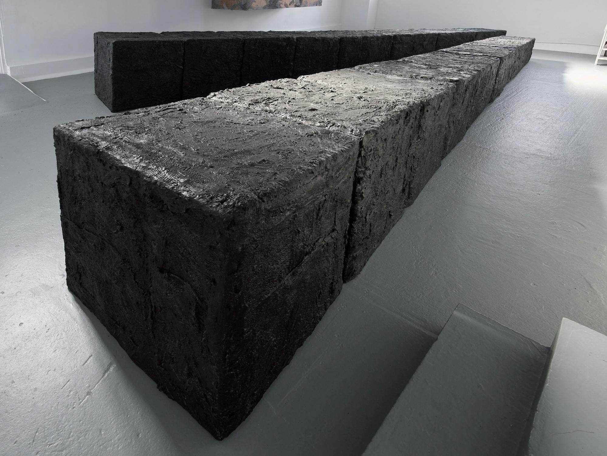 Two rows of large, rough-textured, black rectangular blocks are placed parallel on a light grey floor in a minimalist, white-walled room. The blocks resemble compressed charcoal or soil, creating a stark contrast with the clean surroundings.
