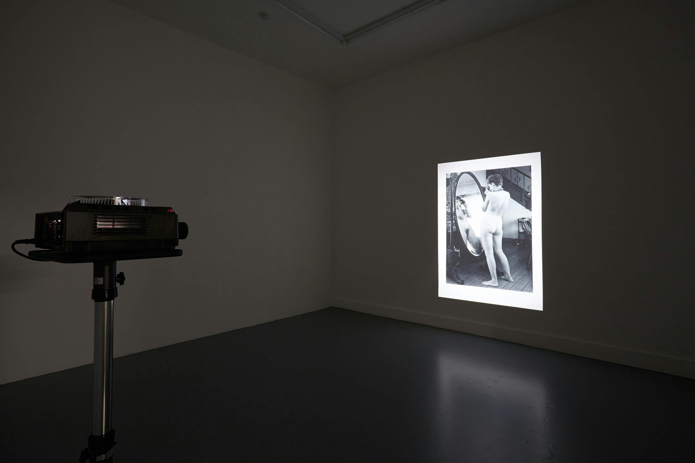 A dimly lit room with a slide projector on a stand projecting a black-and-white image onto a white wall. The image shows a woman standing in front of a mirror, partially draped with a towel. The floor and walls of t