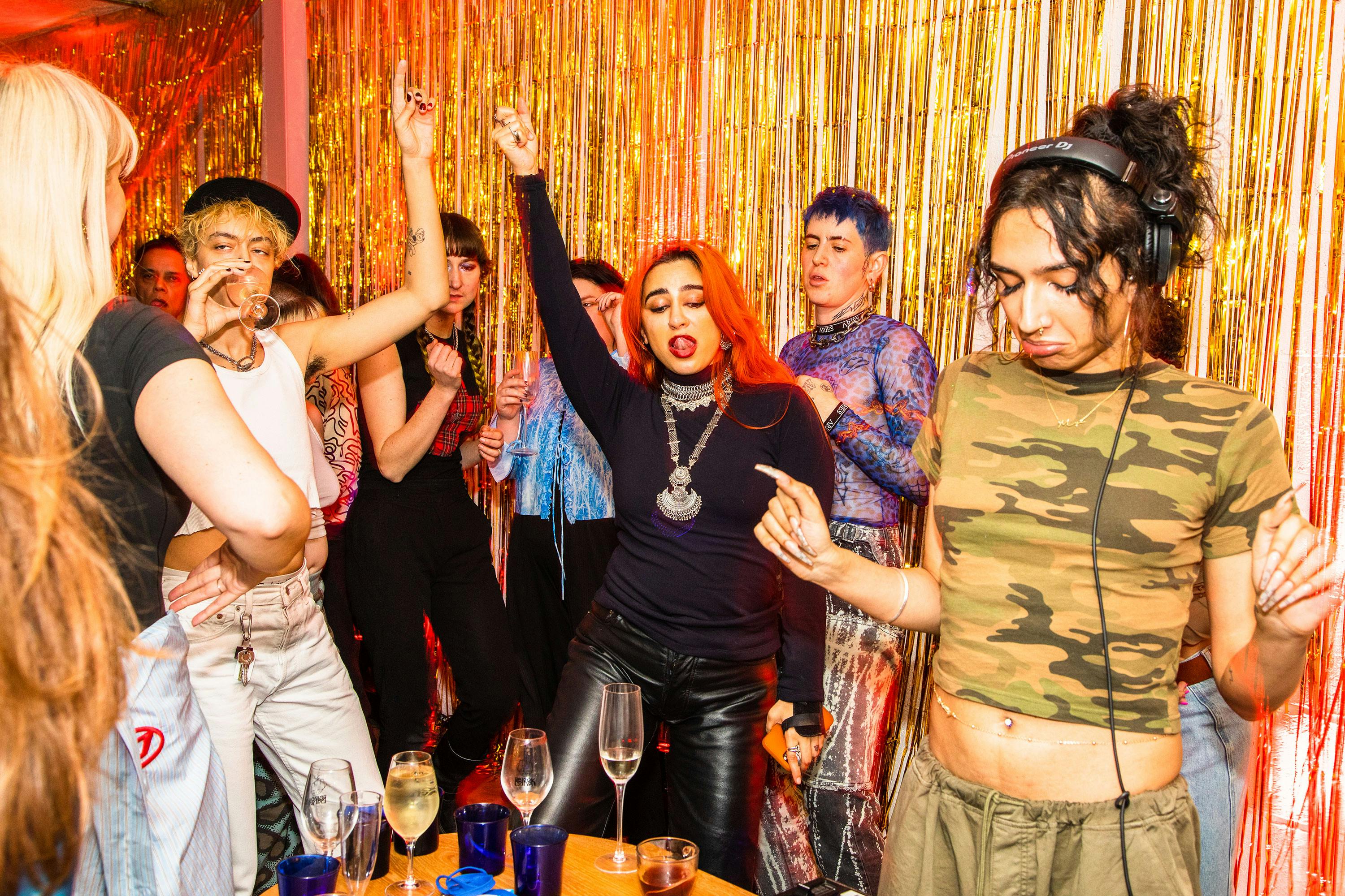 A group of people dancing energetically at a party in front of gold tinsel streamers, with drinks on a table in the foreground. One person in the center poses confidently, and everyone appears to be enjoying the lively atmosphere.
