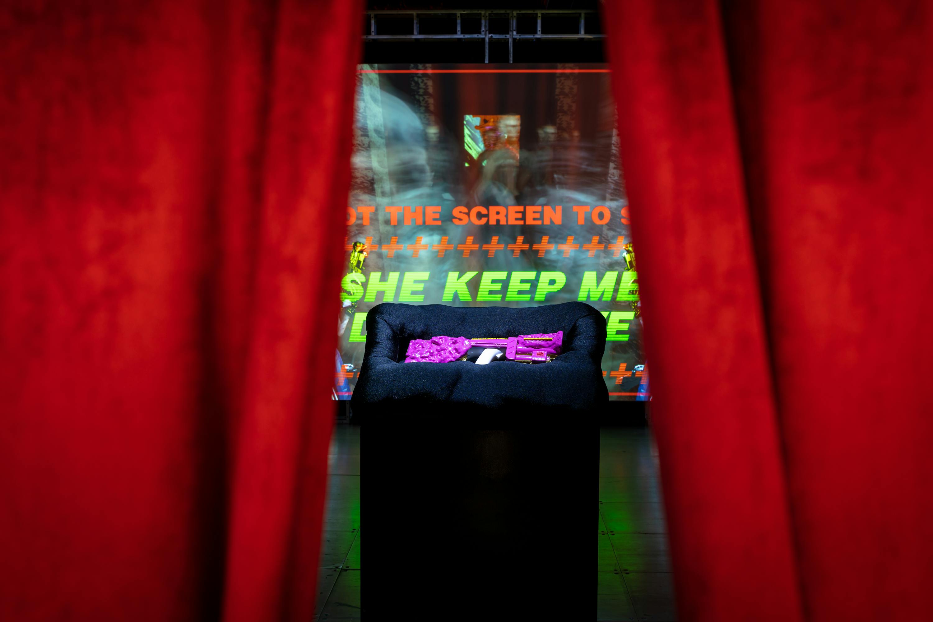 A bright pink machine gun rests on black pillow upon a black plinth, glimpsed between a gap in a red curtain
