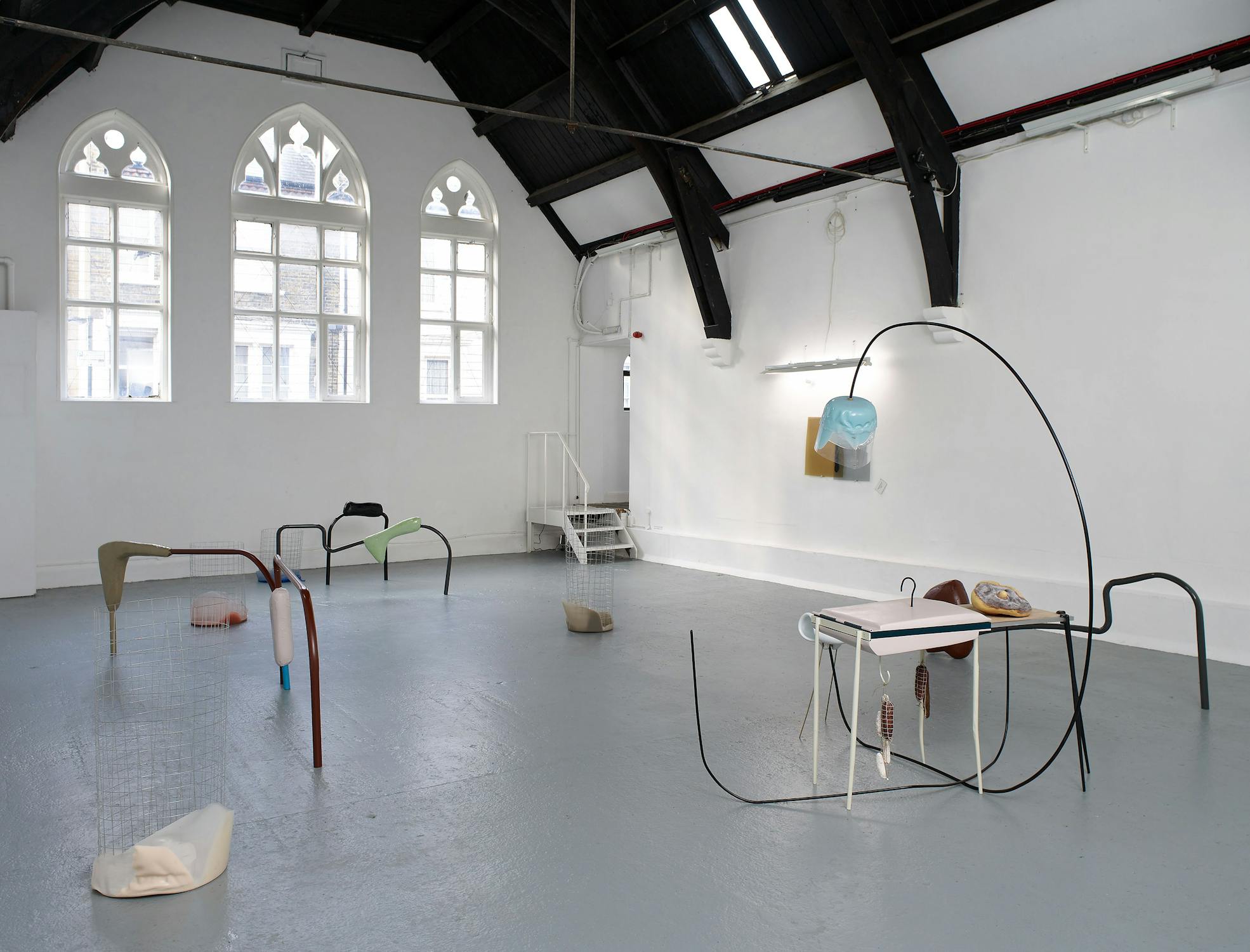 An art gallery with a minimalist installation, featuring abstract sculptures of various shapes and colors placed on the floor. The room has arched windows, exposed beams, and a stairway leading to a small platform. The walls are white and the floor is grey.