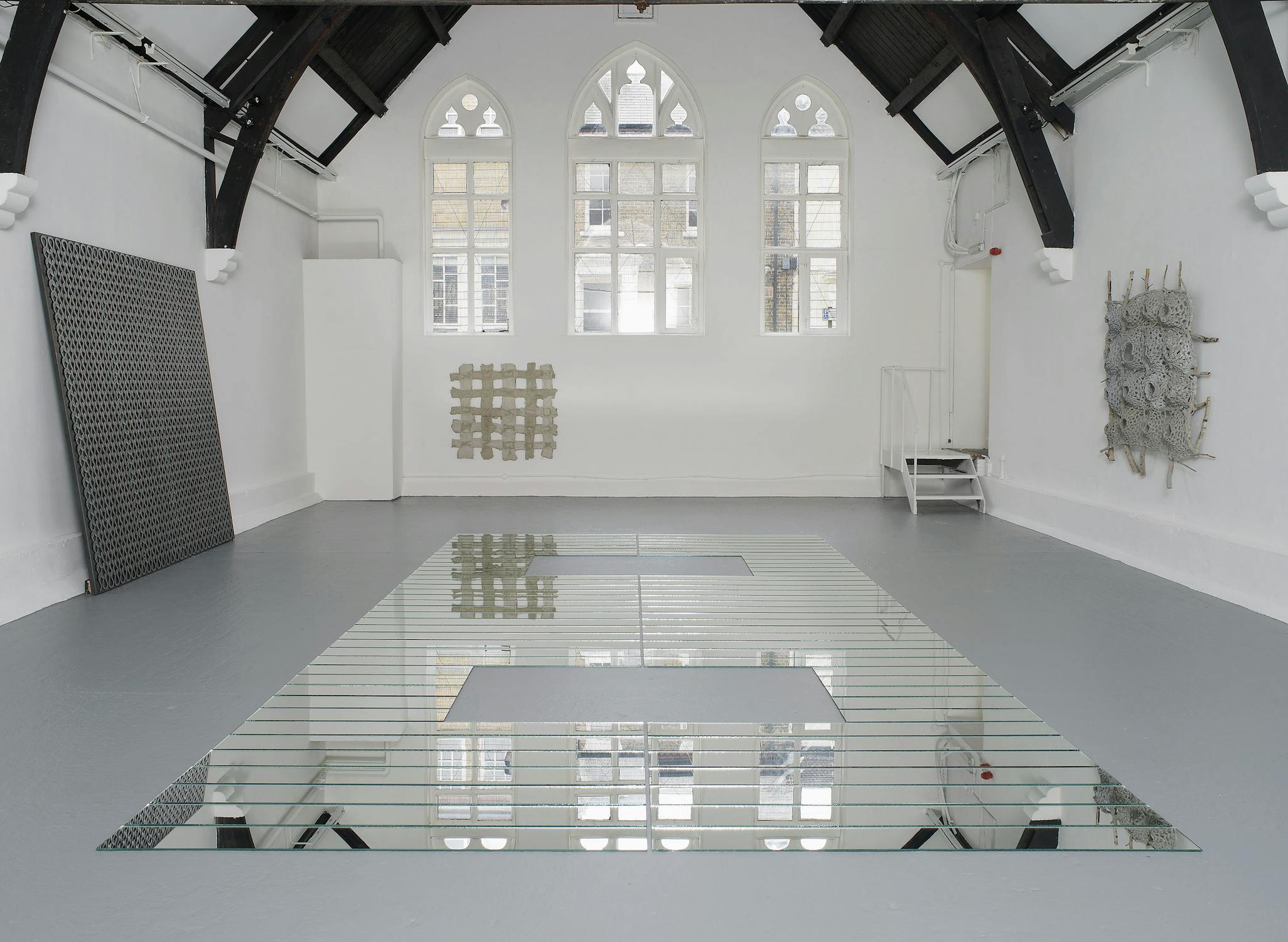 A minimalist gallery with arched windows, white walls, and exposed dark beams. The floor features a large geometric mirrored installation. Three abstract wall sculptures—one metallic, one grid-like, one intricate—are displayed along the walls.