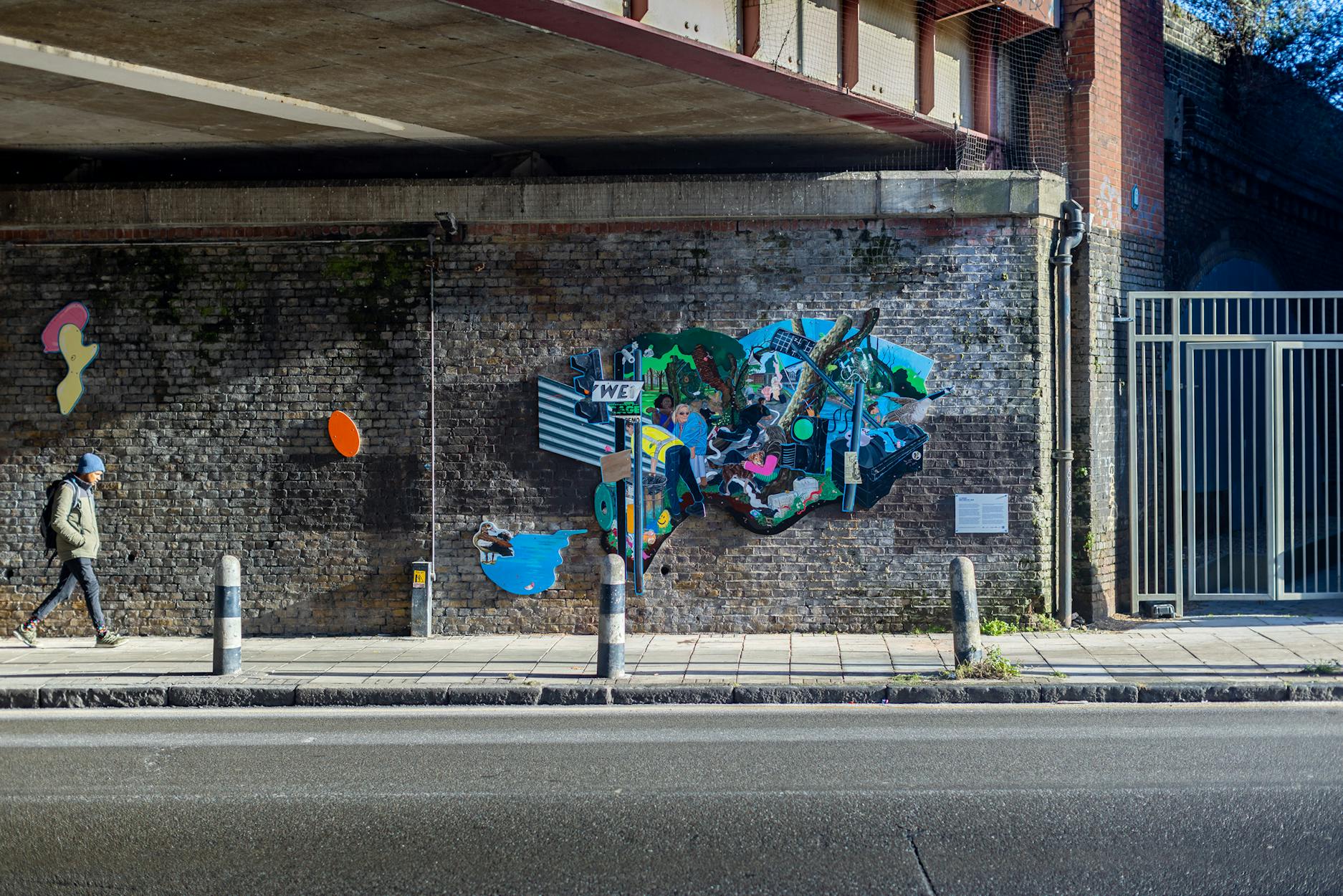 A vibrant and eclectic mural by Flo Brooks is painted on a brick wall. The mural features various elements like a man holding a baby, wildlife such as birds, and urban imagery including street signs and graffiti. There's also a small plaque with text on the wall beside the mural.