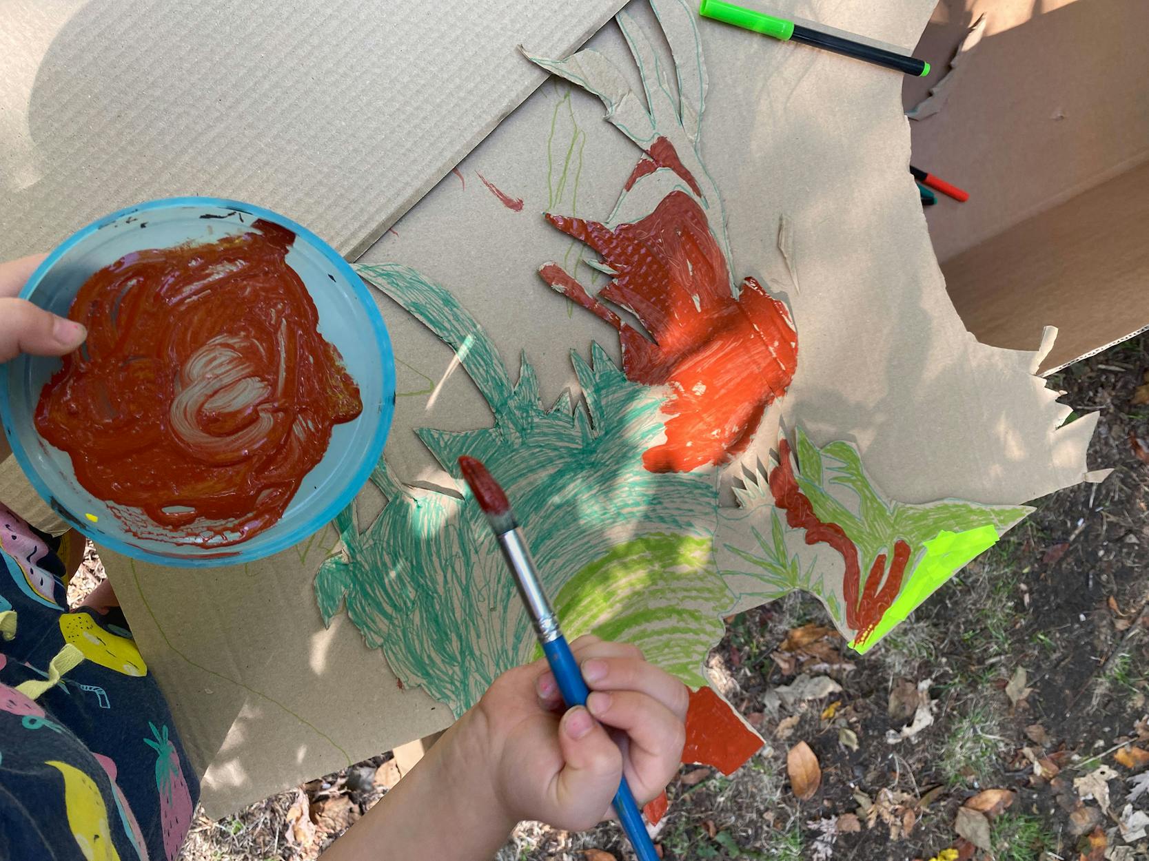 A person is painting on a cardboard with red and green paint, using a brush. The cardboard is on the ground outdoors, surrounded by leaves. A blue bowl with red paint is held in one hand. Some markers are also visible.