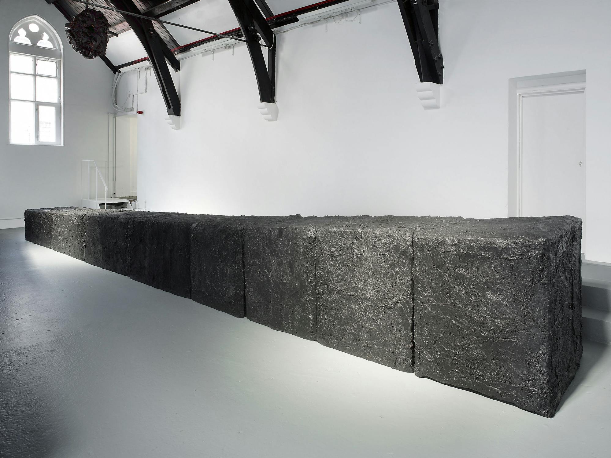 A long, rectangular sculpture made of rough, dark, stone-like blocks sits in a white-walled gallery with high ceilings and exposed beams. Light from large windows and spotlights highlights the texture of the blocks. A door and small staircase are in the background.