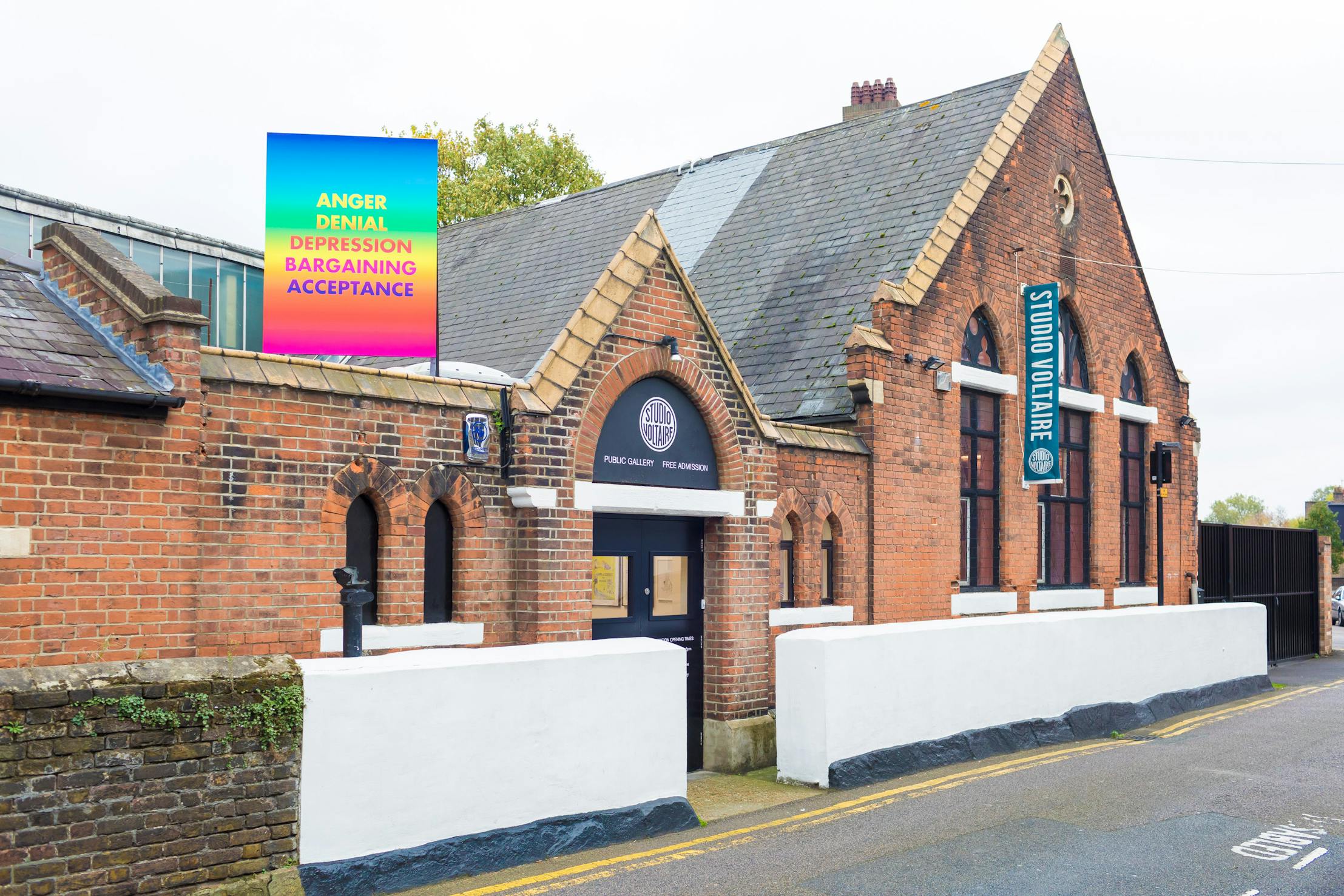 Studio Voltaire's gallery building with peaked roofs and arched windows has a sign reading "Studio Voltaire." A colourful billboard on the roof features a rainbow gradient with the words: ANGER, DENIAL, DEPRESSION, BARGAINING, ACCEPTANCE.