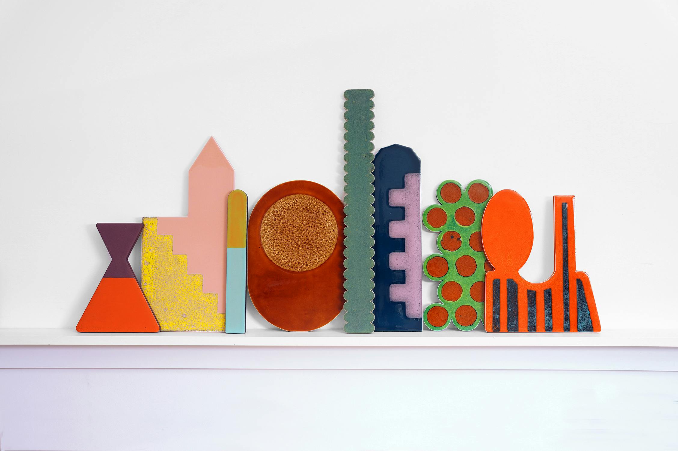 A collection of colorful geometric tiles by Lubna Chowdhary resembling abstract buildings arranged in a row against a white wall. Shapes include triangles, circles, and rectangles, featuring a mix of textures and patterns in vibrant colors.