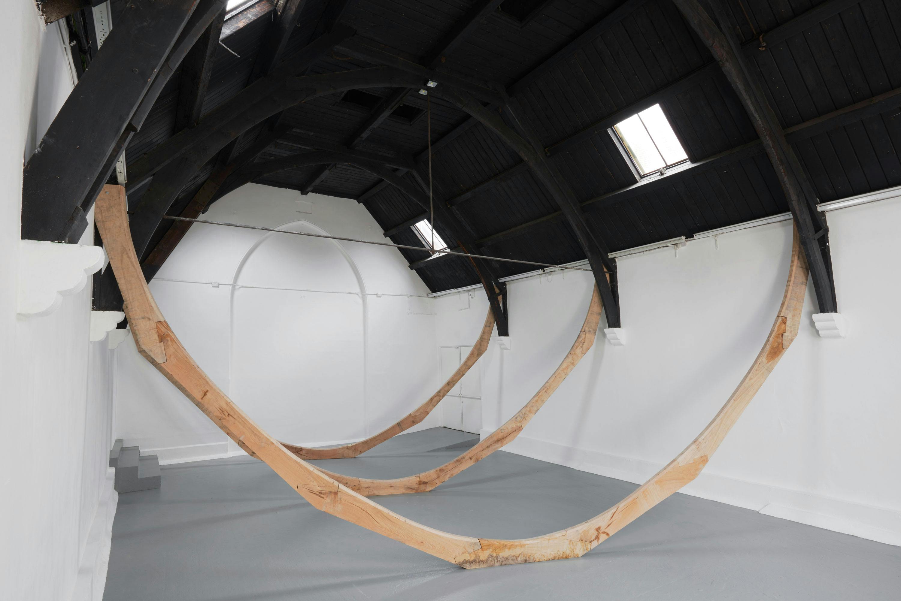 A minimalist gallery with white walls and a high, dark wooden ceiling features two large, curved wooden beams suspended from the ceiling, forming intersecting arcs that dominate the empty space. Light enters through skylights, illuminating the installation.
